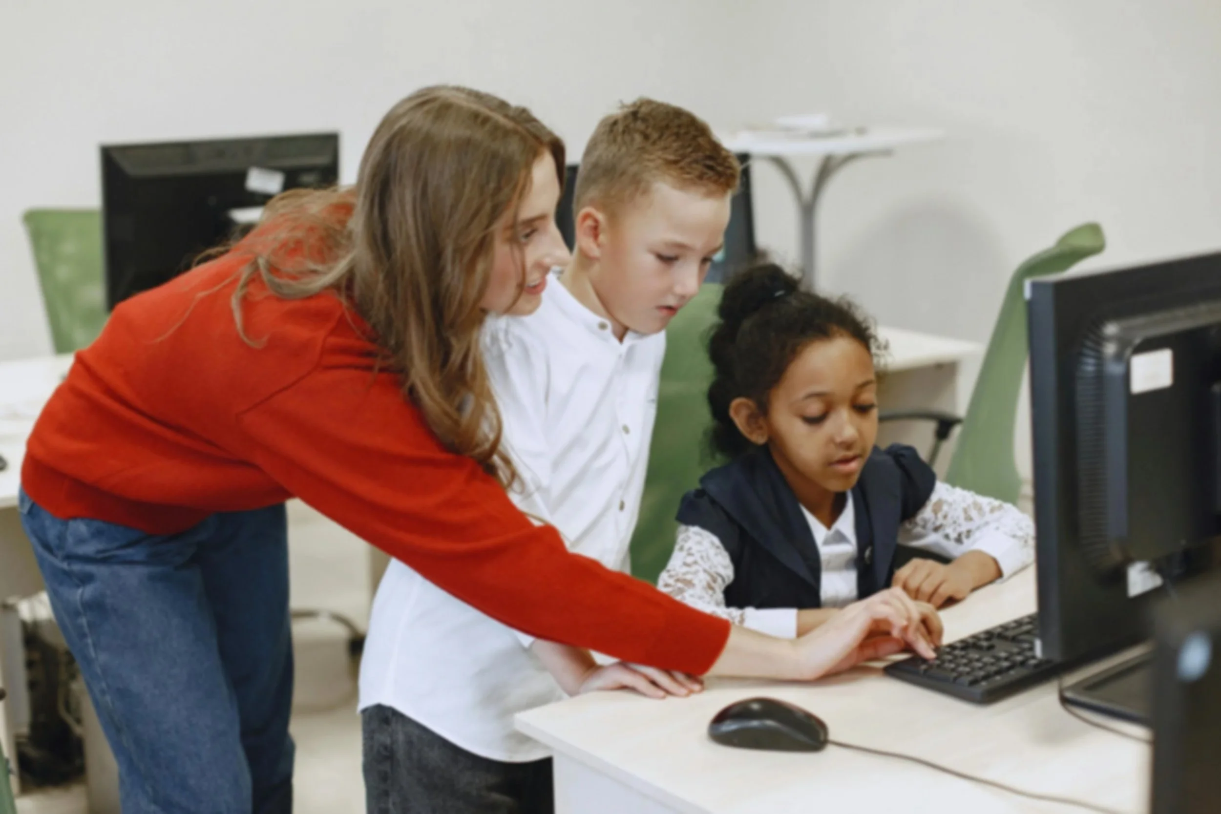 Teacher helping two elementary students use a desktop computer during a digital learning activity in a classroom.