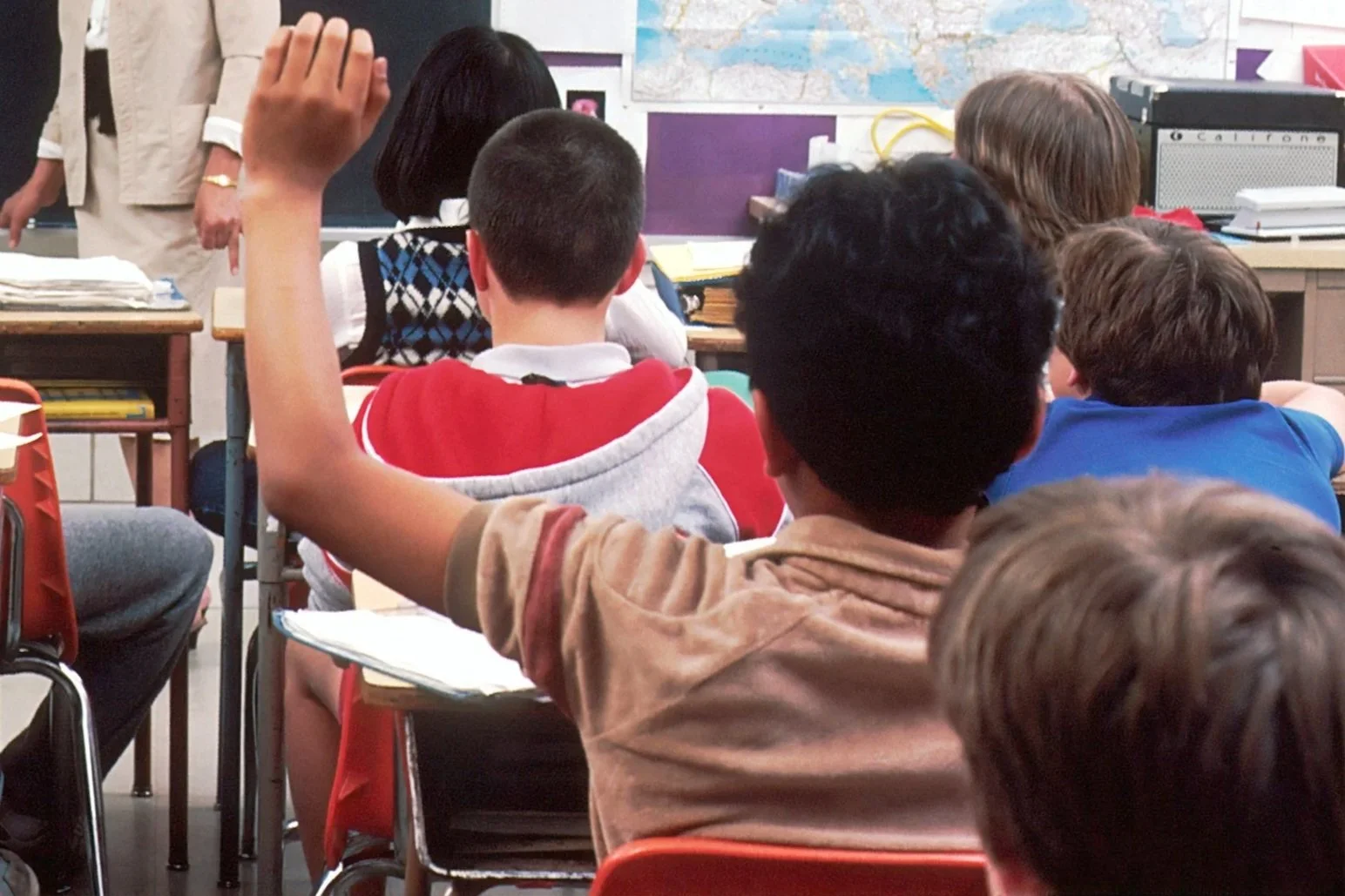 Student raising hand in a classroom while other students sit facing the teacher during a lesson.