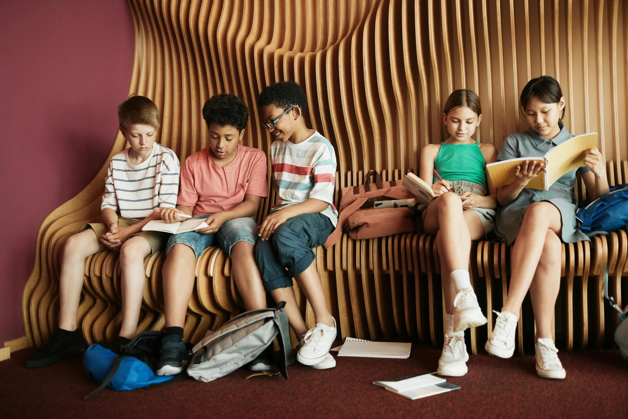 Group of diverse students sitting together on curved wooden seating while writing in notebooks and working on classroom assignments.