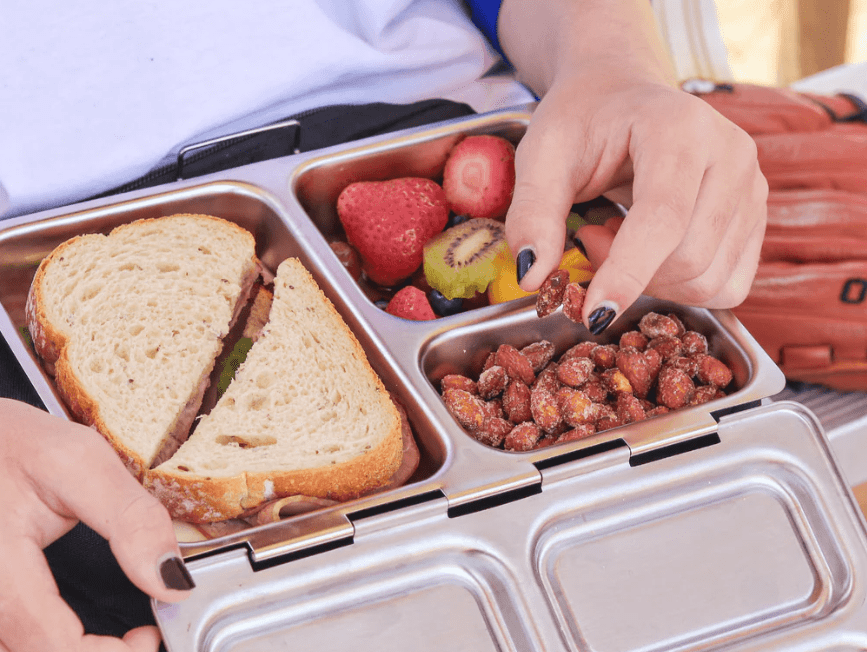 A student holding an open PlanetBox lunchbox filled with a sandwich, fresh fruit, and roasted nuts, demonstrating portable meal convenience.