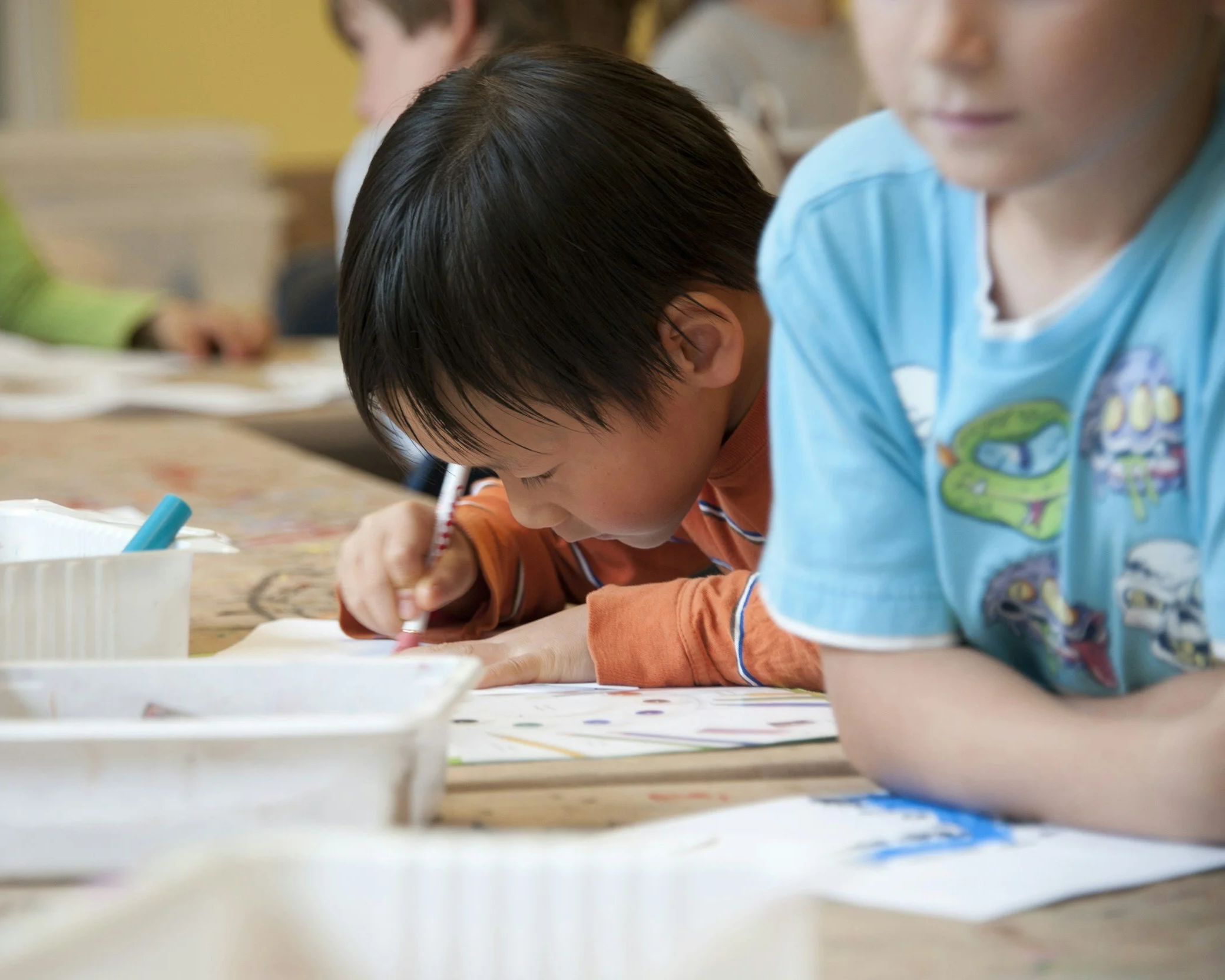 Young student focused on writing or drawing at a classroom table, with other children and art materials visible nearby.