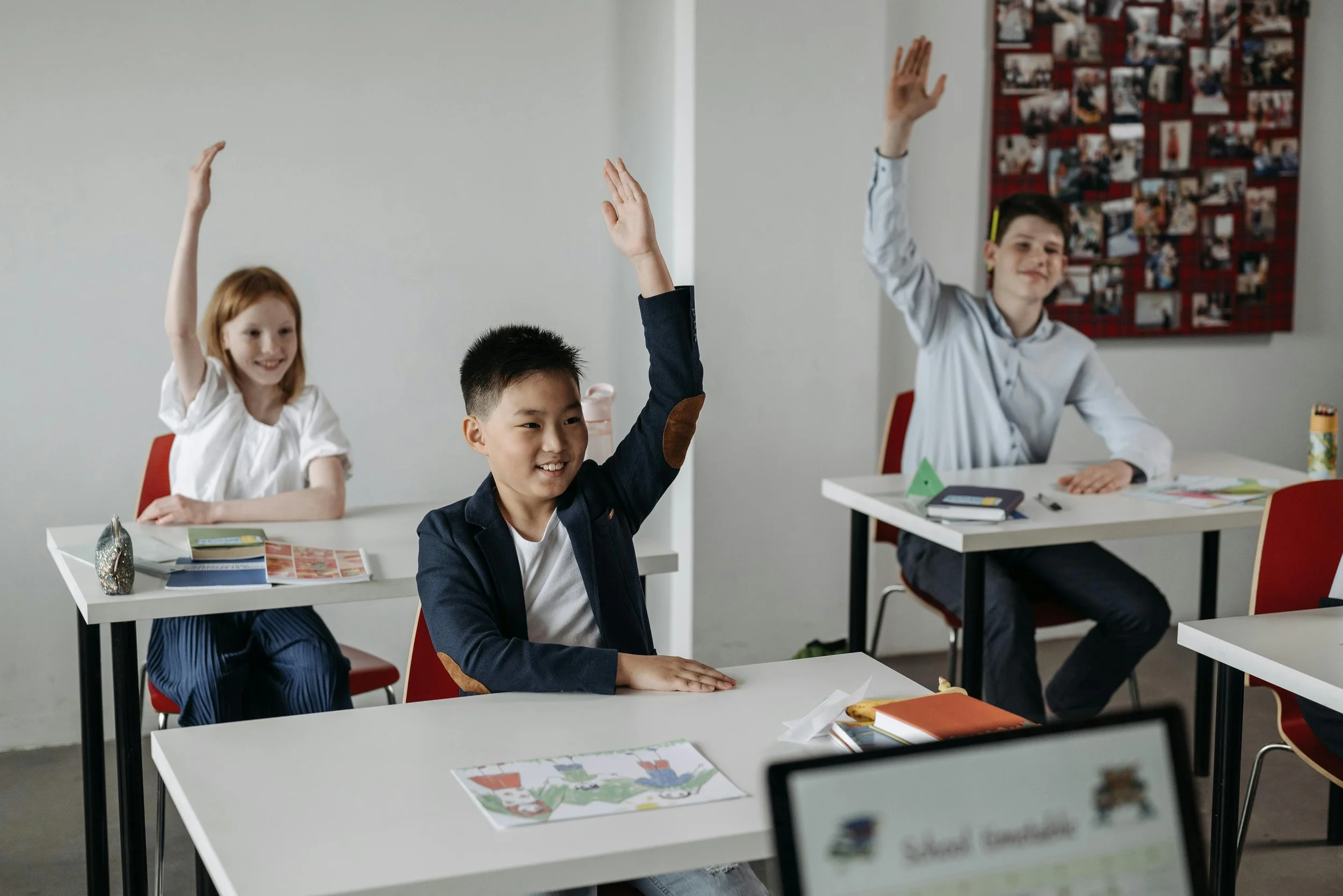 Middle school students in a classroom seated at desks with their hands raised, actively participating in a lesson.