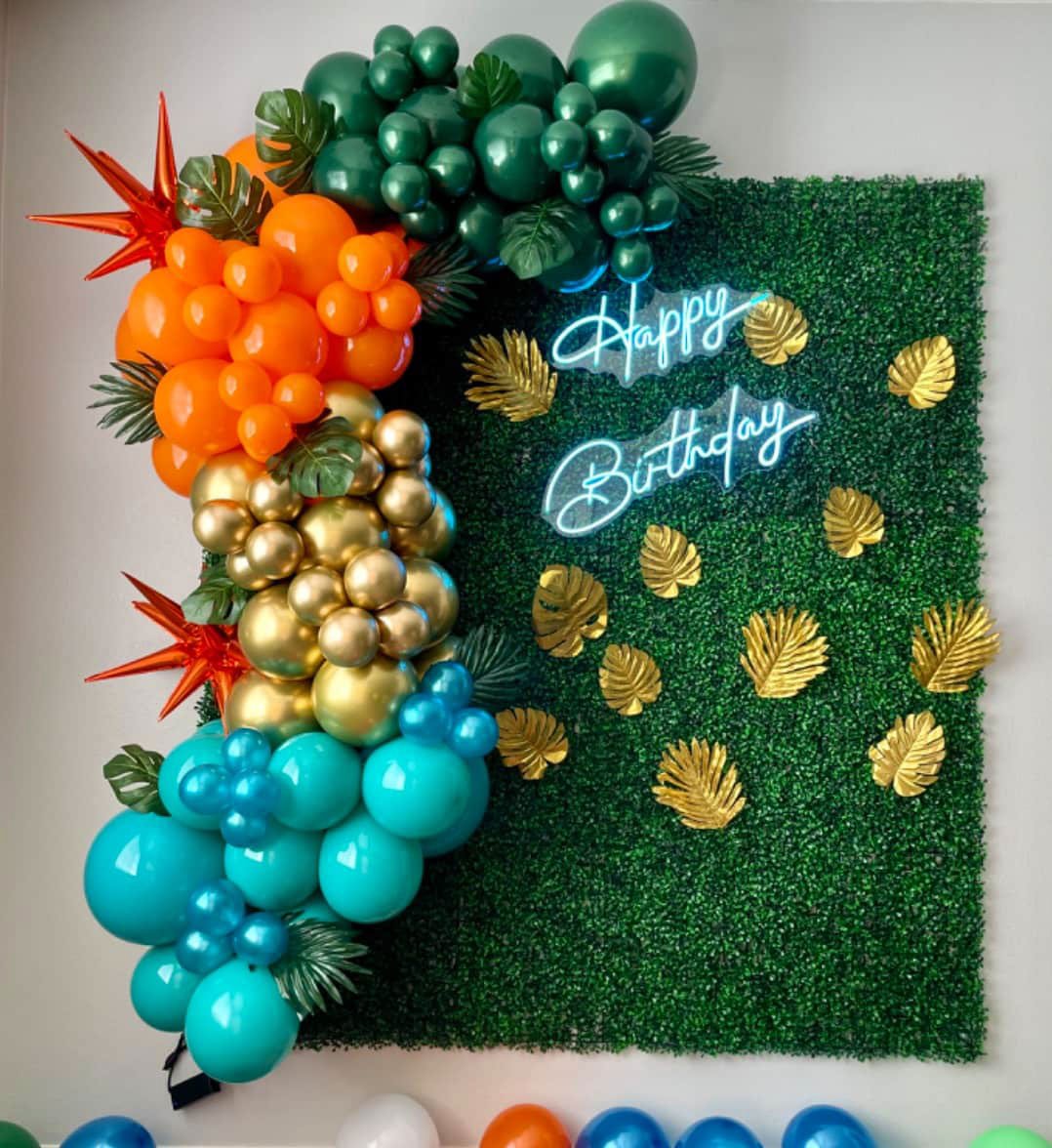 Colorful balloon arch and green backdrop with "Happy Birthday" neon sign and gold leaf decorations.