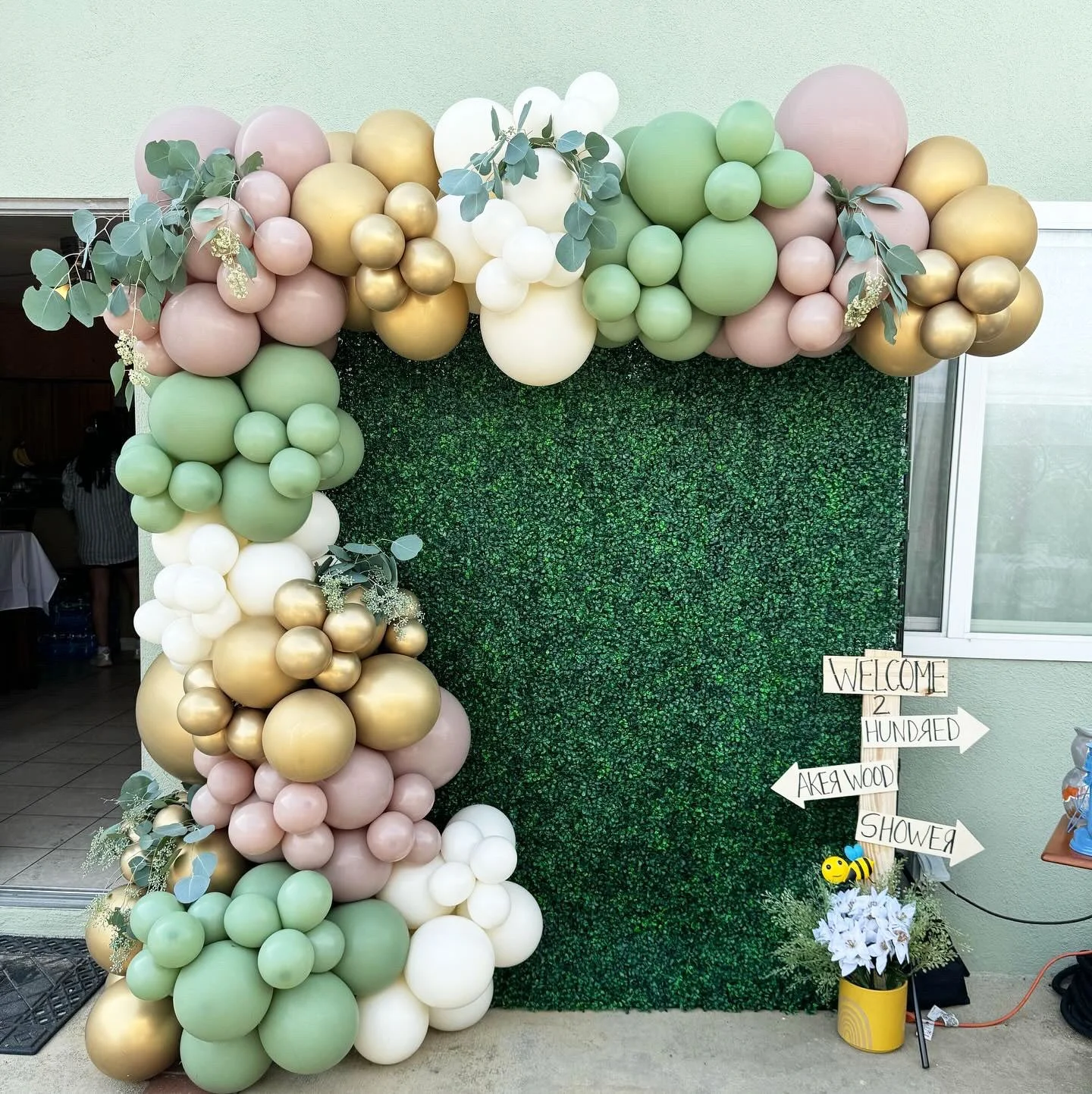 A decorative backdrop featuring clusters of balloons in green, gold, white, and pink arranged on a green foliage wall. Eucalyptus leaves accentuate the balloon arrangement. There is a wooden sign nearby with handwritten text and a pot with white flow