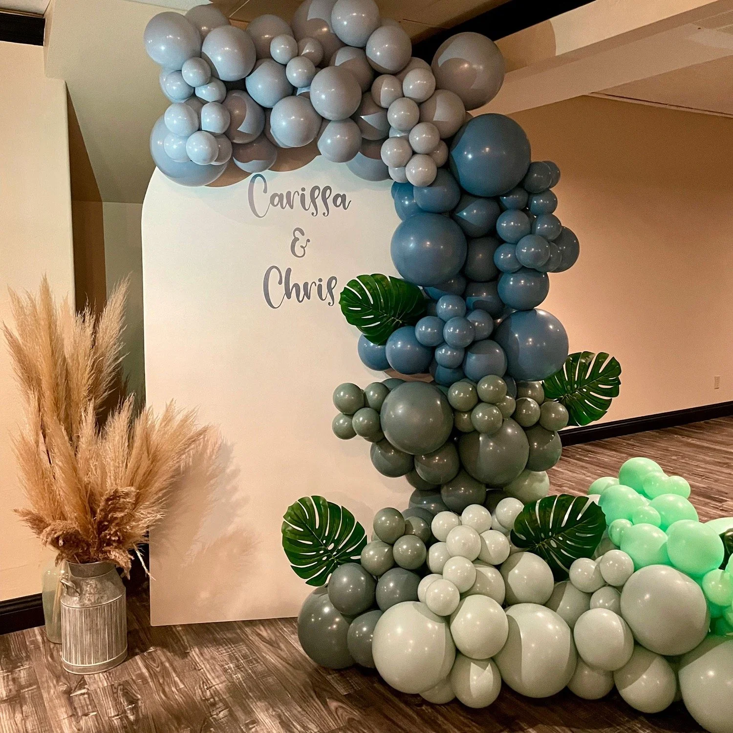 Balloon arch decoration with blue and mint green balloons, featuring green leaves, next to a sign reading 'Carissa & Chris' and pampas grass in a vase on a wooden floor.