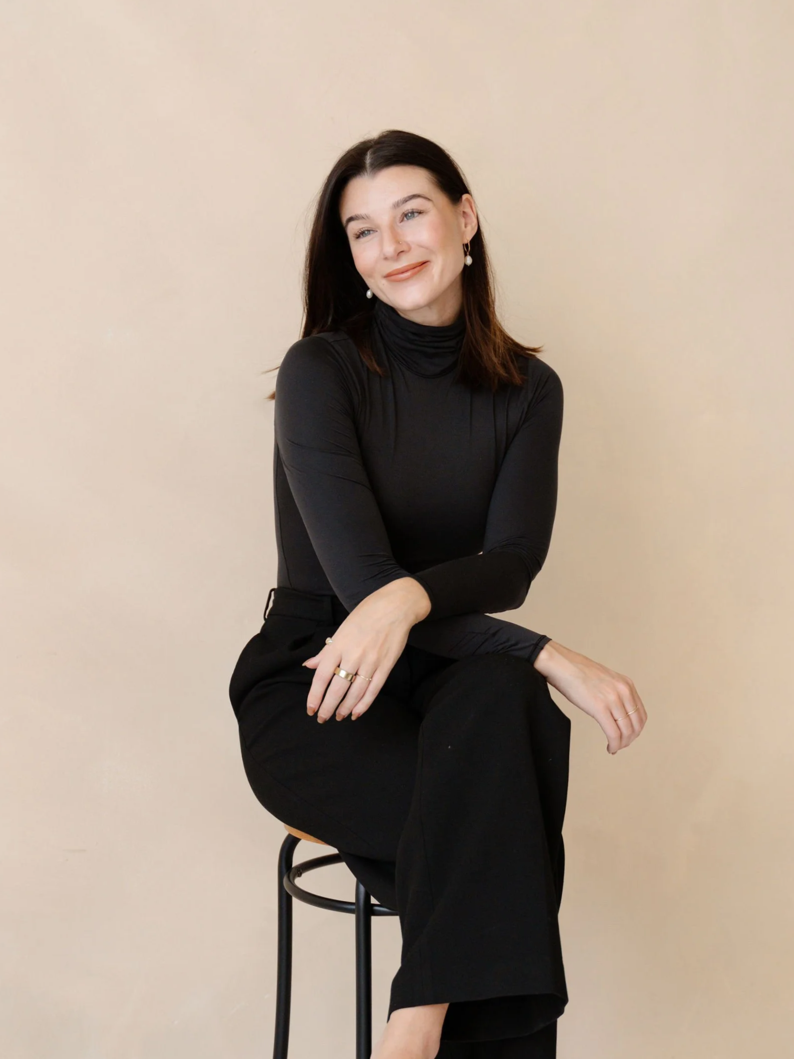 A woman with dark hair wearing a black turtleneck and black pants, sitting on a stool against a beige background, smiling with her arms crossed.