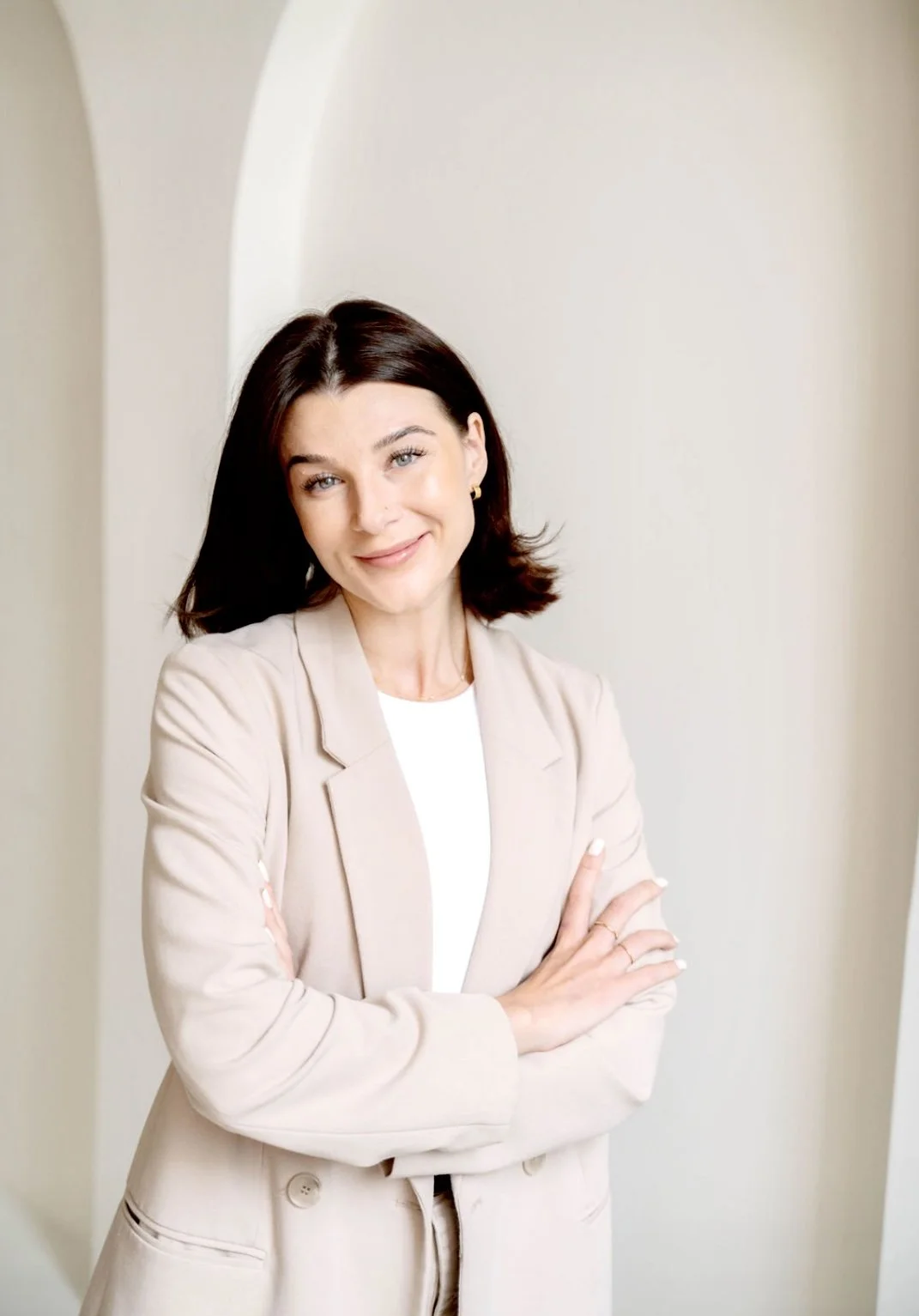 A woman with dark brown hair, wearing a beige blazer and white shirt, smiling with crossed arms in front of a light-colored background.