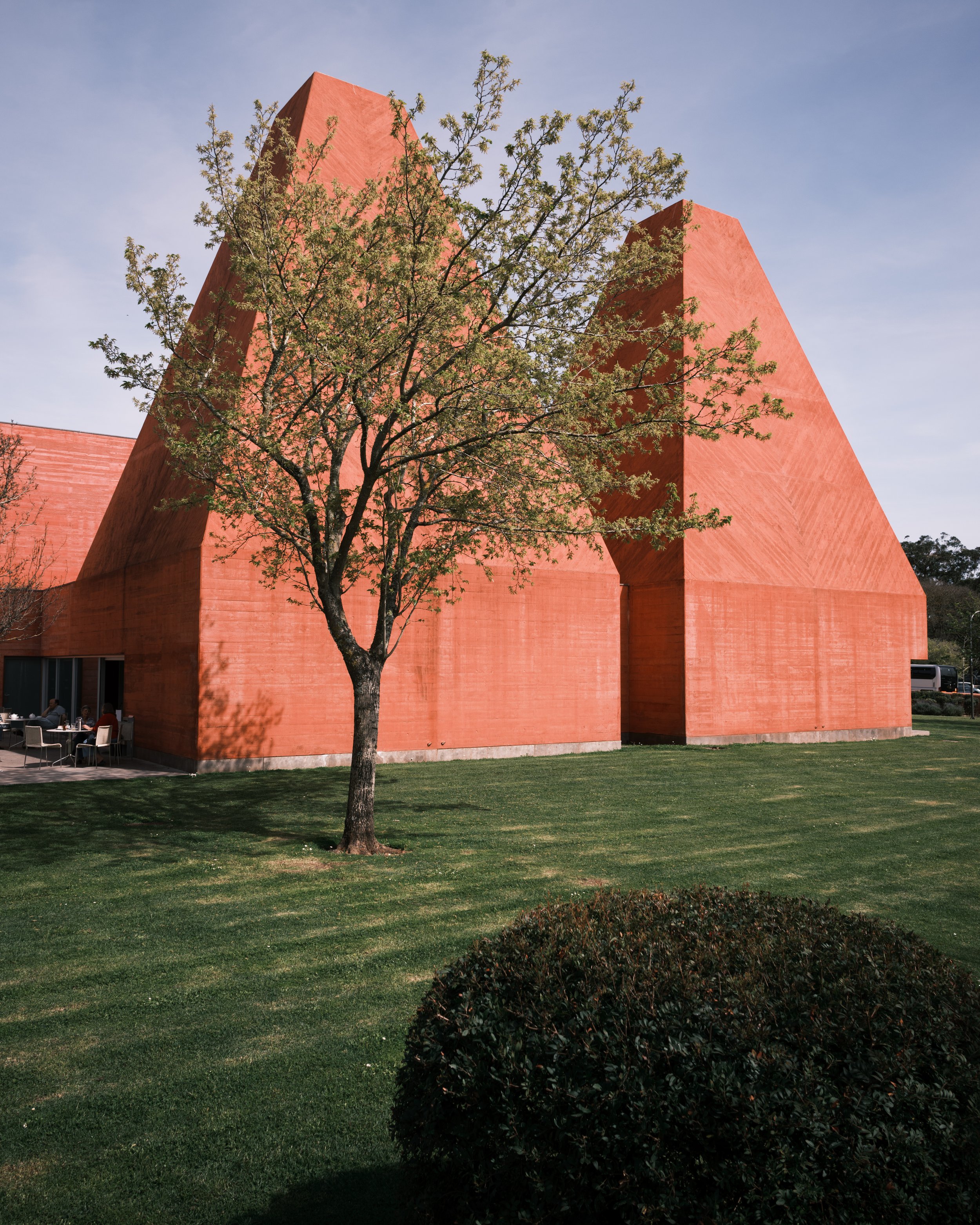 Modern red concrete building with angular shapes, surrounded by green lawn and trees.