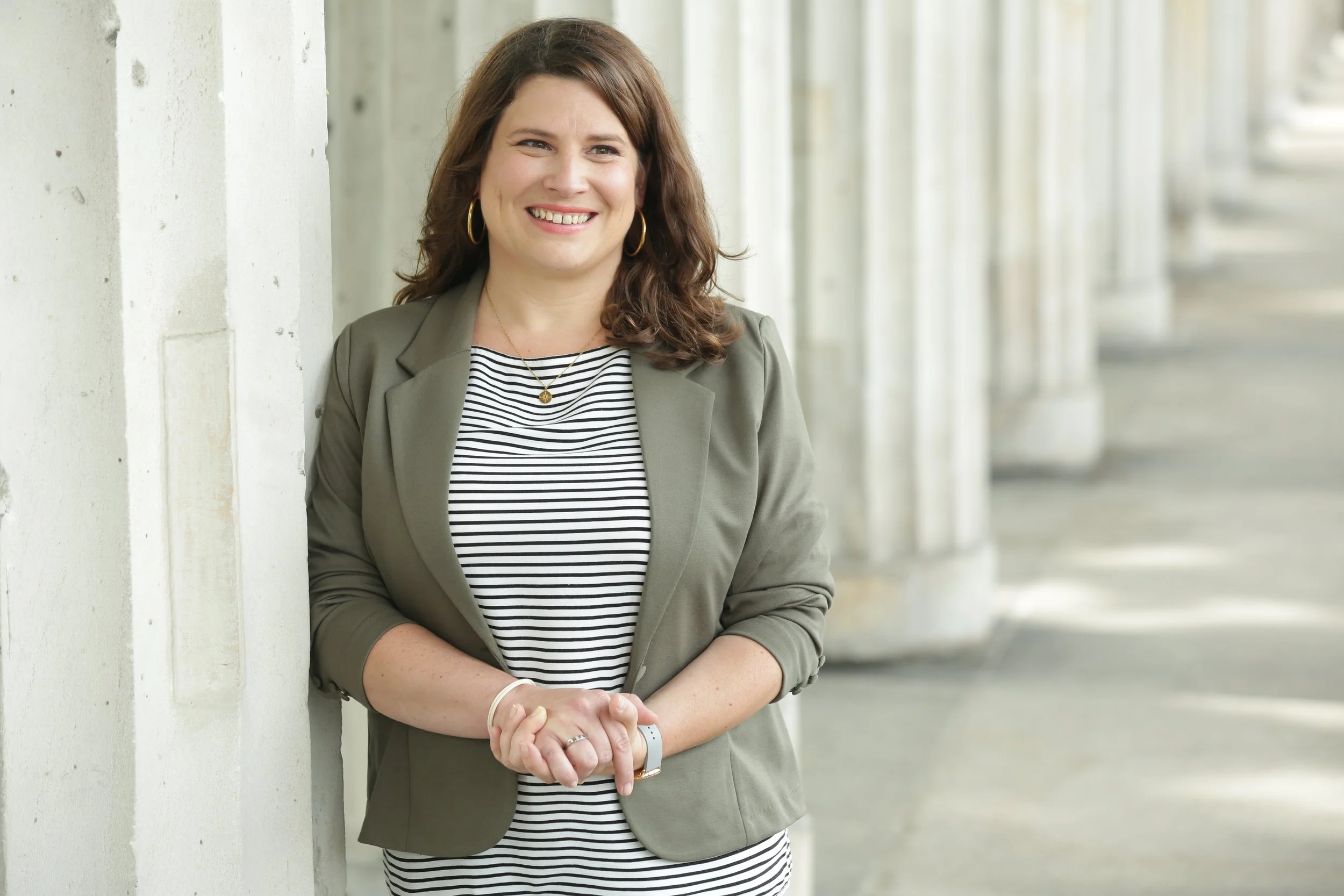 Frau in gestreiftem Kleid und olivegrünem Blazer lehnt an einer Säule und lächelt.