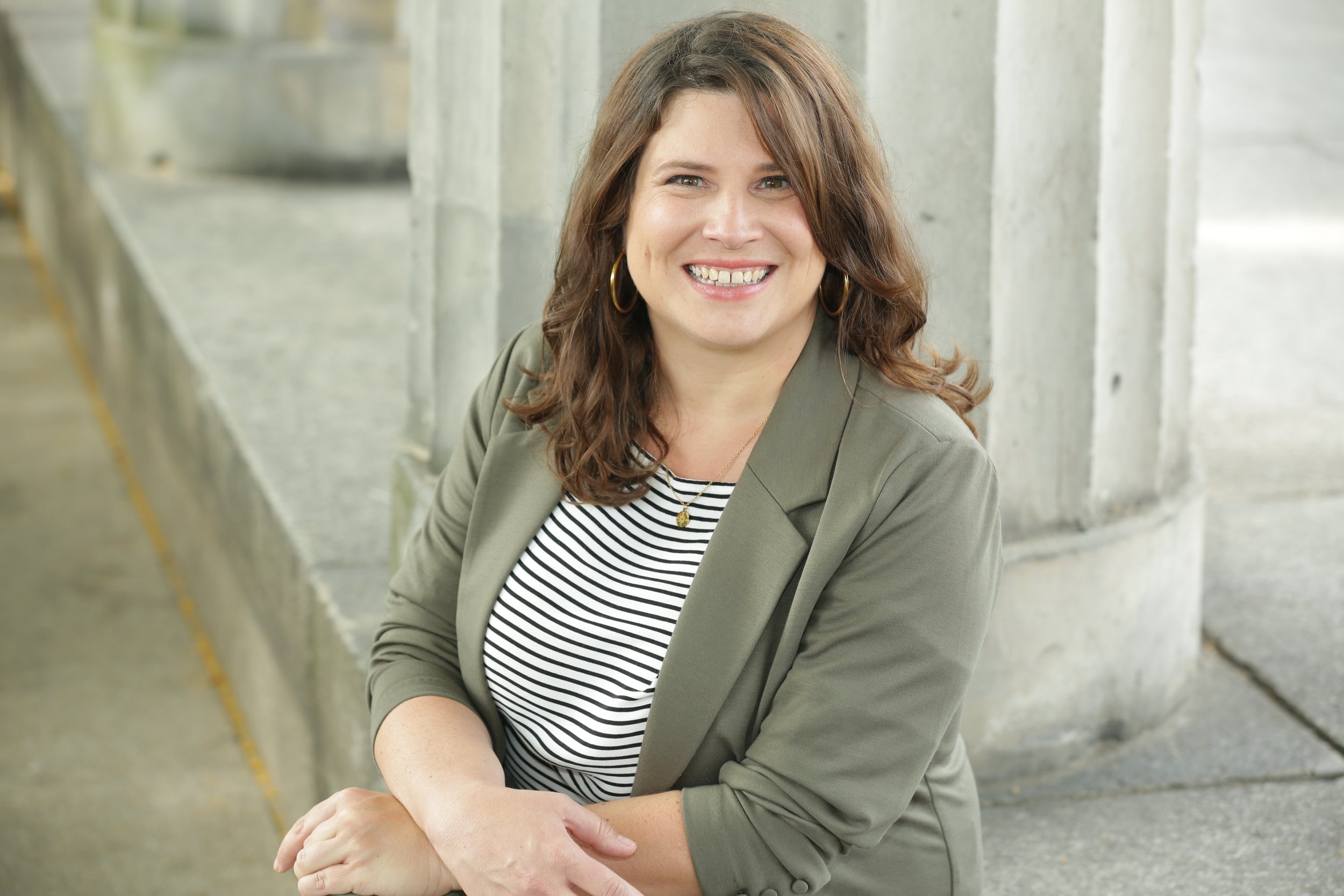 Frau mit braunem Haar in gestreiftem Shirt und grünem Blazer lächelt, lehnt sich an eine Betonstruktur.