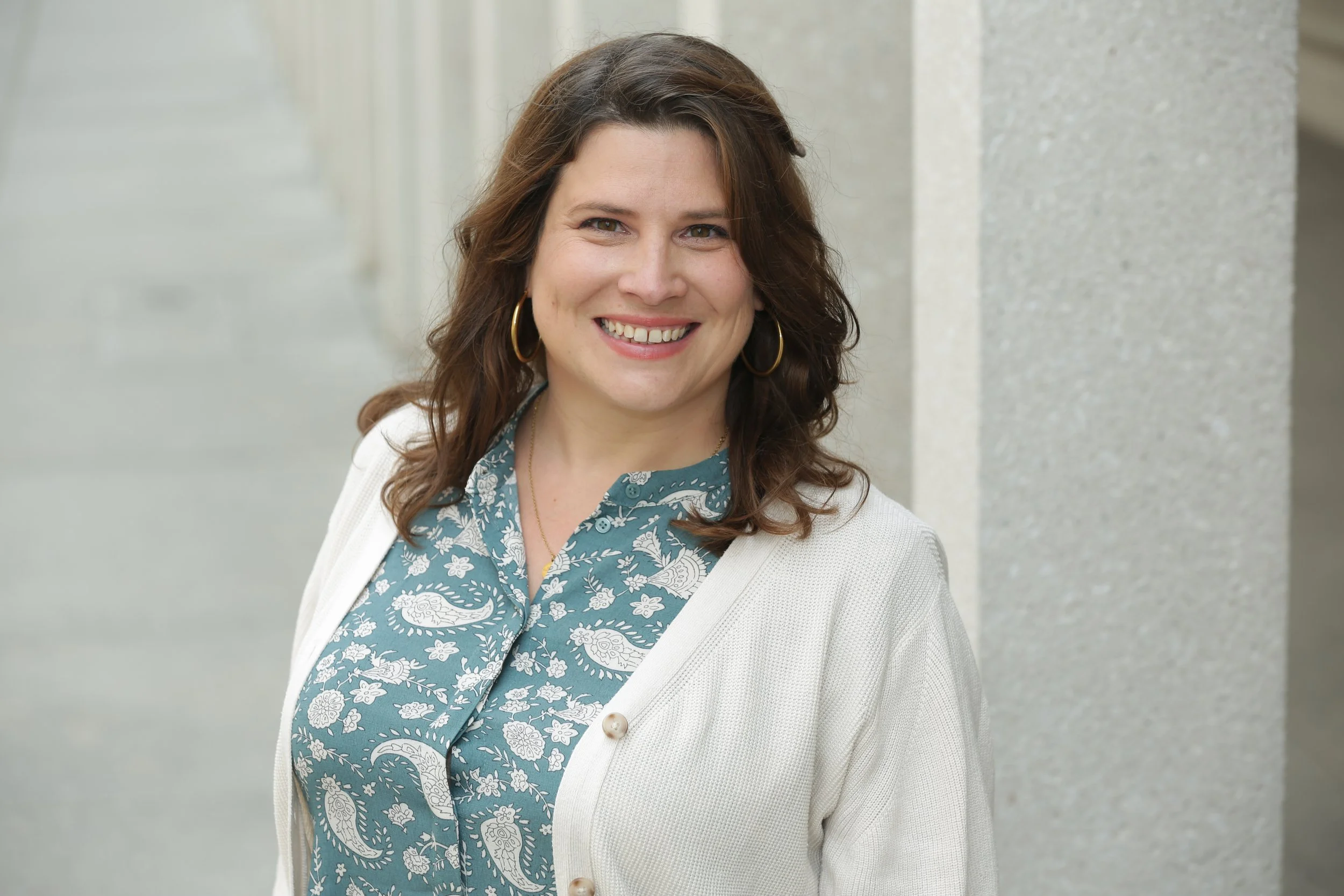 Frau mit blauer gemusterter Bluse und weißer Strickjacke steht lächelnd vor grauem Hintergrund.