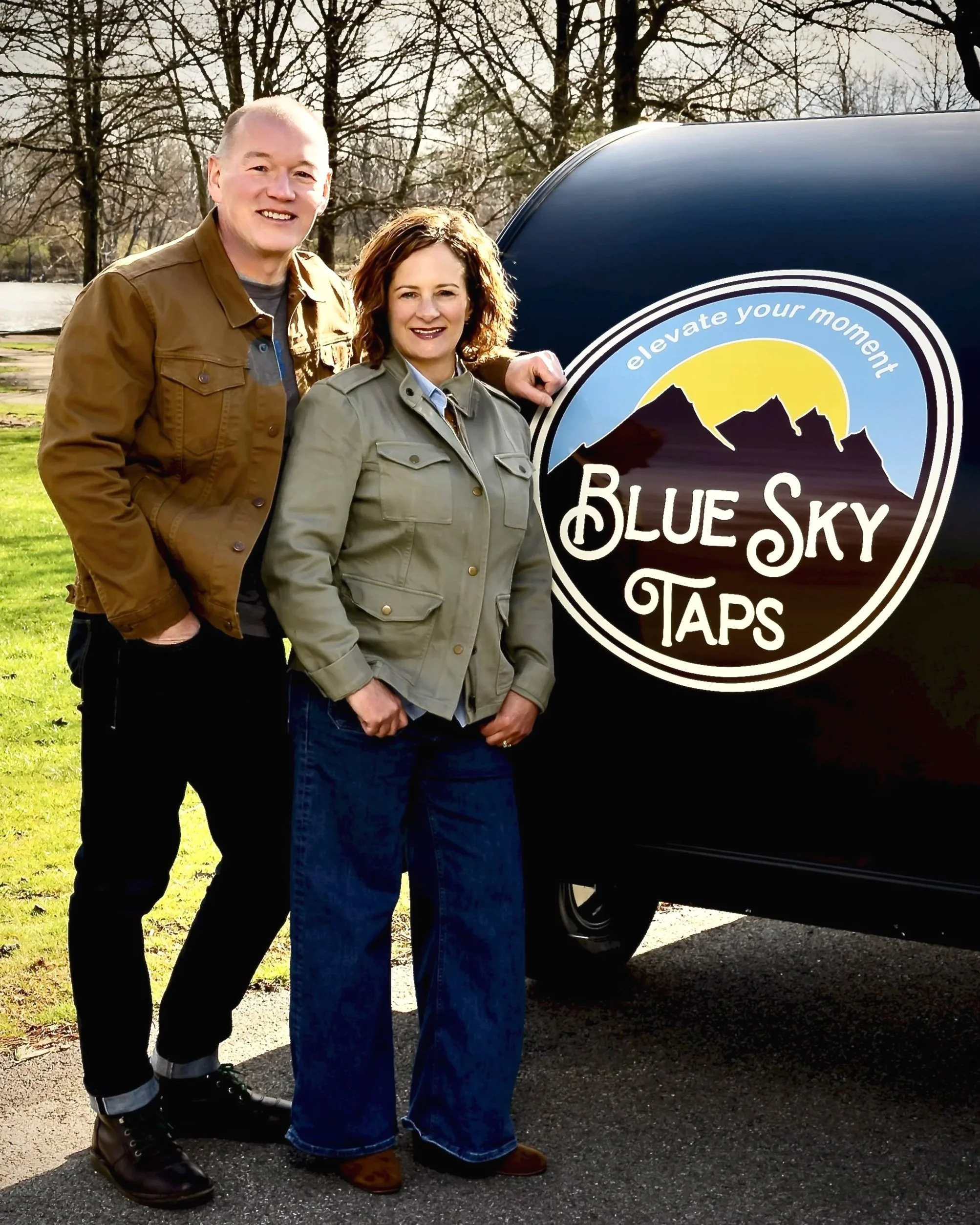 A smiling man and woman standing outdoors on a sunny day in front of the Blue Sky Taps mobile bar and tap trailer.  The scene features trees and a natural park setting.