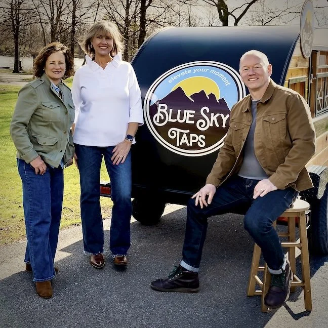 Three bar tenders, two women and one man, standing outdoors near the Blue Sky Taps mobile tap trailer and bar in a park-like setting with trees in the background at party in Columbus, Ohio.