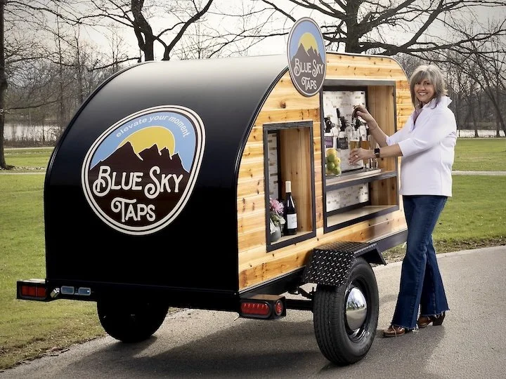 Woman in a white jacket and blue jeans pouring from a tap at a mobile bar trailer labeled 'Blue Sky Taps', parked on a park path near a lake and trees at a wedding in Columbus, Ohio..