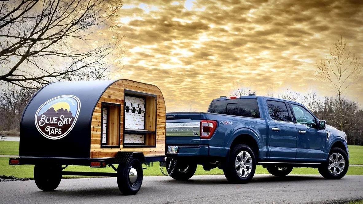 Blue pickup truck towing a mobile tap trailer with a sign that reads "Blue Sky Taps" parked in park under a pretty day with warm colored clouds.