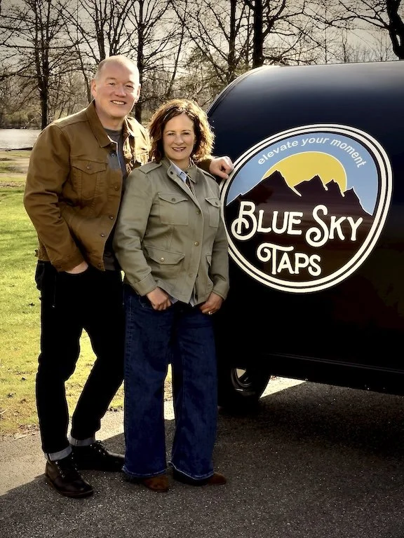 A smiling man and woman standing outdoors on a sunny day in front of the Blue Sky Taps mobile bar and tap trailer and bar service at a wedding in Columbus, Ohio.  The scene features trees and a natural park setting.
