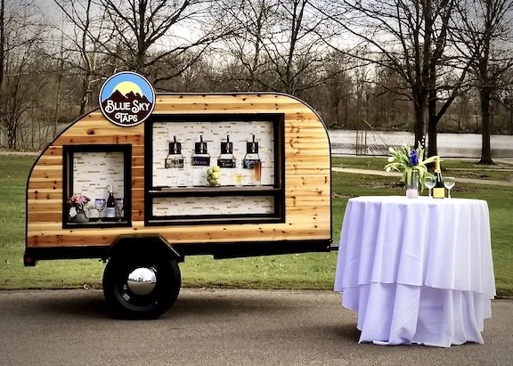 Blue Sky Taps mobile tap trailer and bar setup with decorated table with floral arrangements outdoors near a river and trees.