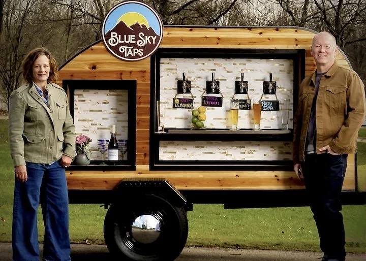 Blue Sky Taps mobile tap trailer and bar service with two people standing on either side, outdoors on grass, under a beautiful sky, and bar setup with drinks, lemons, and flowers in Columbus, Ohio..