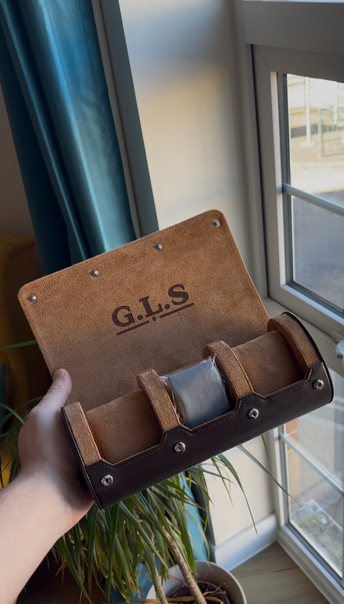 A person holding an open brown leather watch roll with the initials G.I.S printed on the inside flap near a window with a blue curtain and a plant visible.