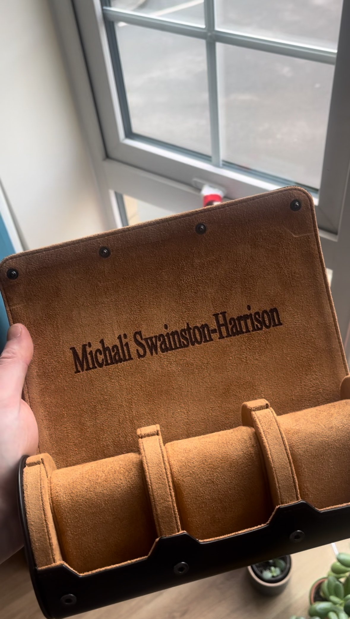 Person holding an open suede watch roll displaying three compartments and a name embossed on the inside cover. A window and potted plant are in the background.