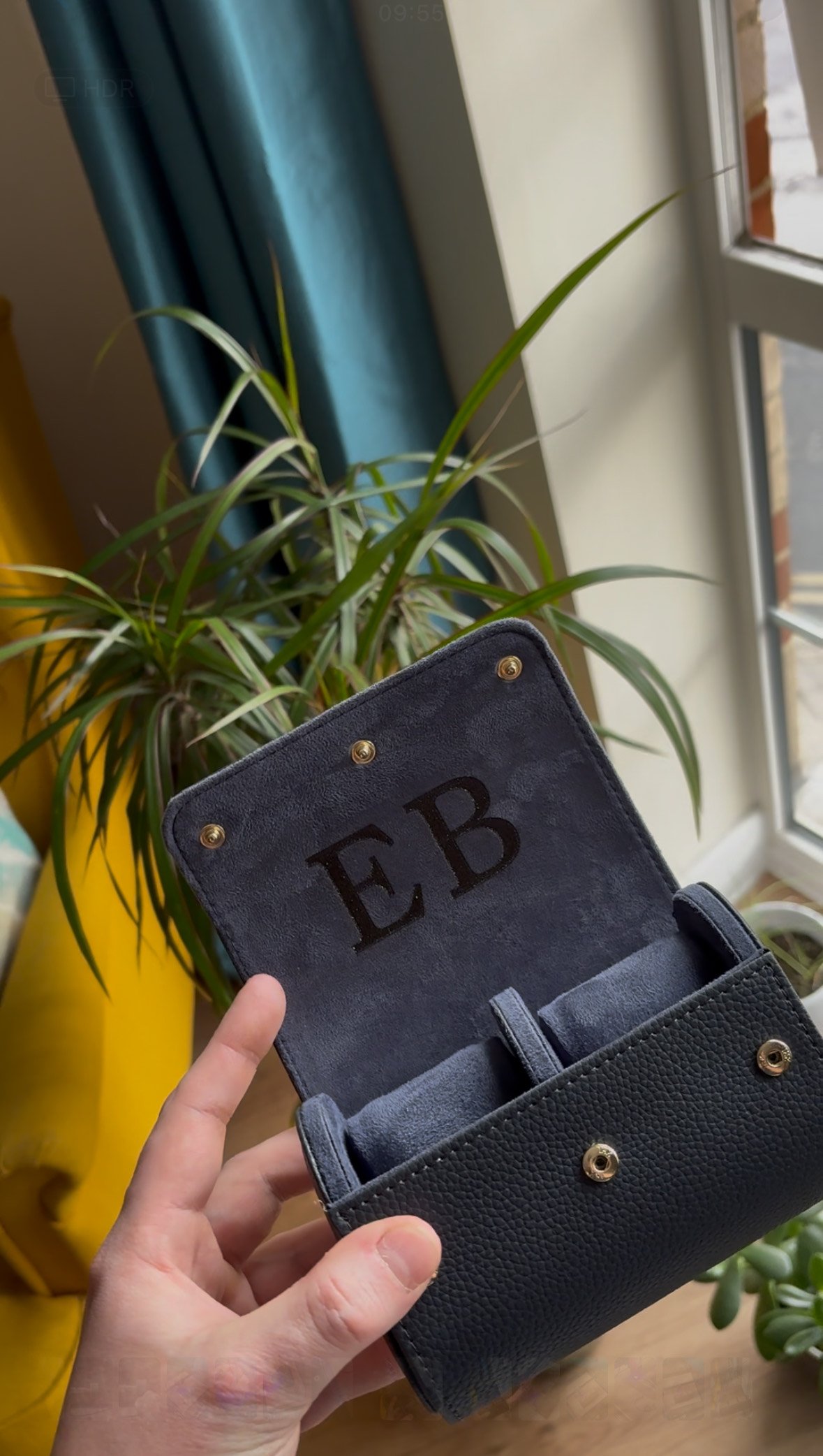 Person holding a black leather pouch with initials 'EB' inside, near a window with green plants in the background.