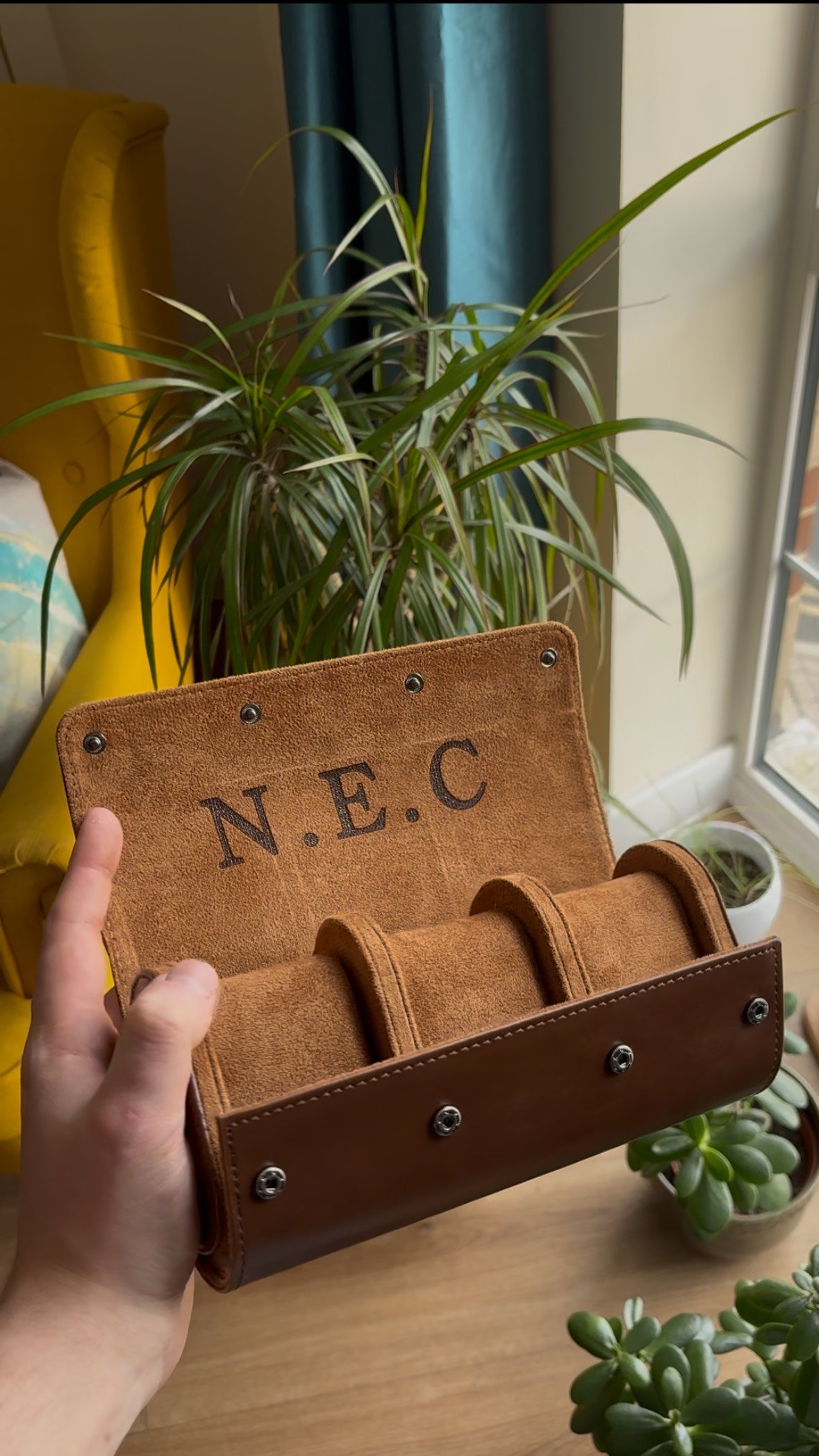Brown leather watch roll with 'N.E.C' initials, held by a hand, surrounded by indoor plants.