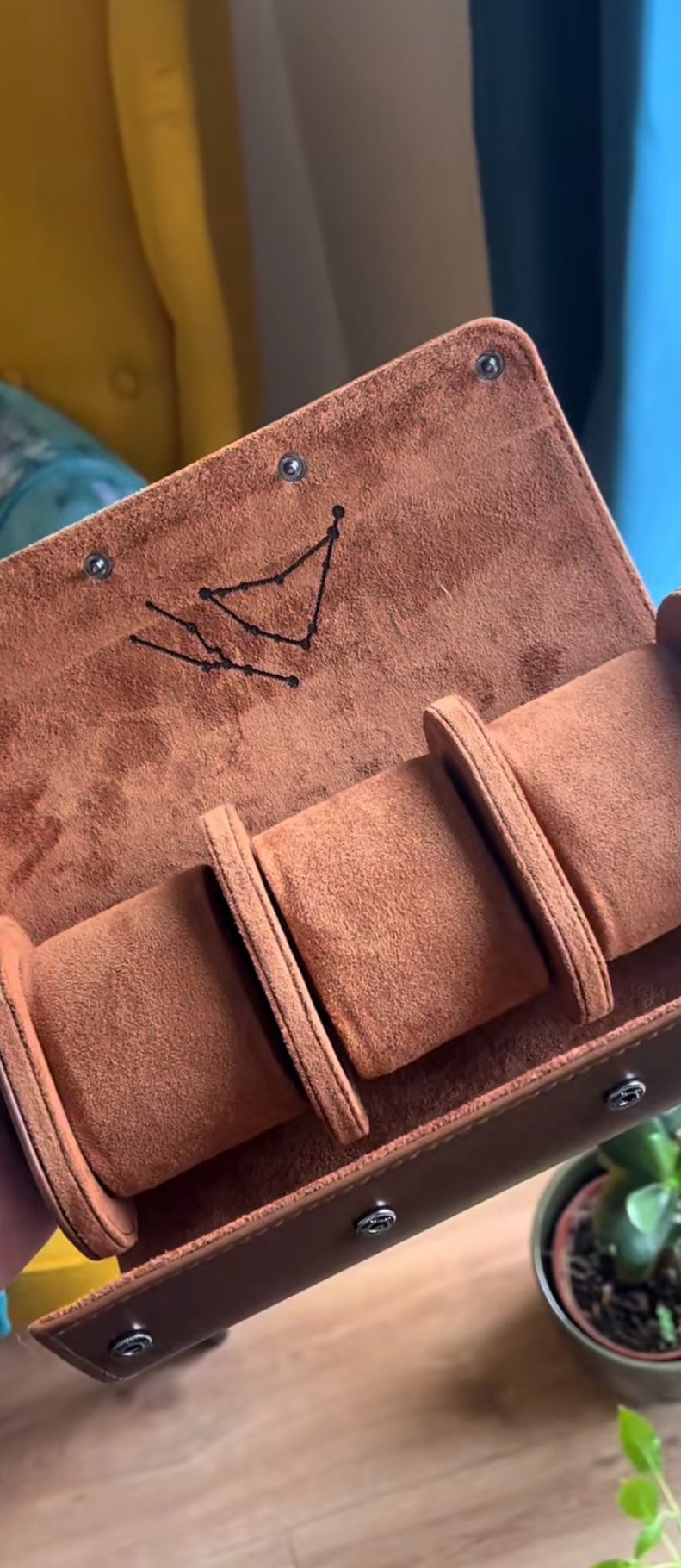 Open brown suede jewelry roll with compartments, embroidery visible inside the flap, and a small potted plant in the background on wooden floor.