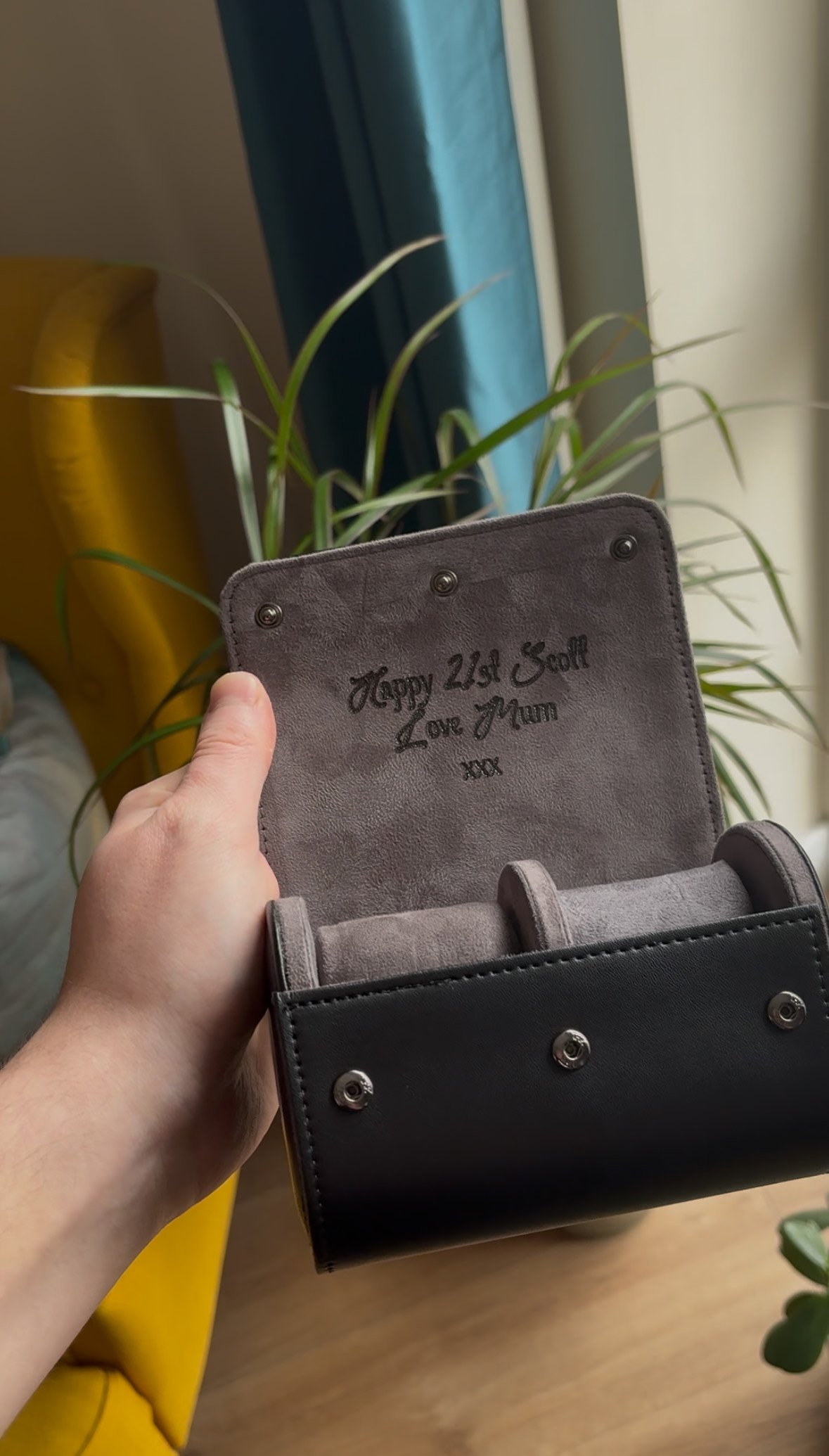 Person holding an open black leather watch case with a personalized message, 'Happy 21st Scott Love Mum xxx,' inside, next to a houseplant and a yellow chair.