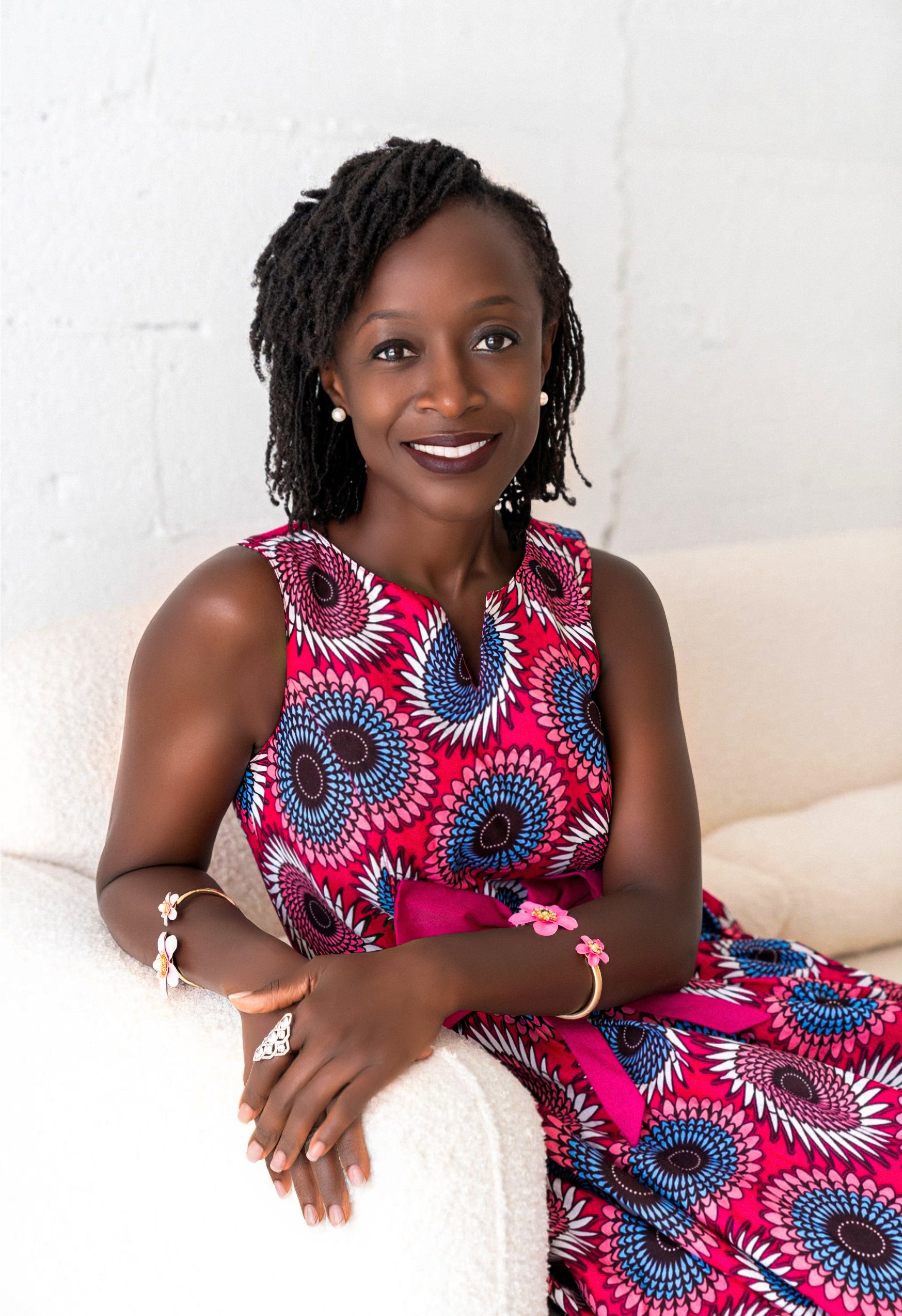 A woman with dark skin and shoulder-length dreadlocks wearing a vibrant pink, blue, white, and black patterned dress, sitting on a white couch, smiling at the camera, against a white wall background.