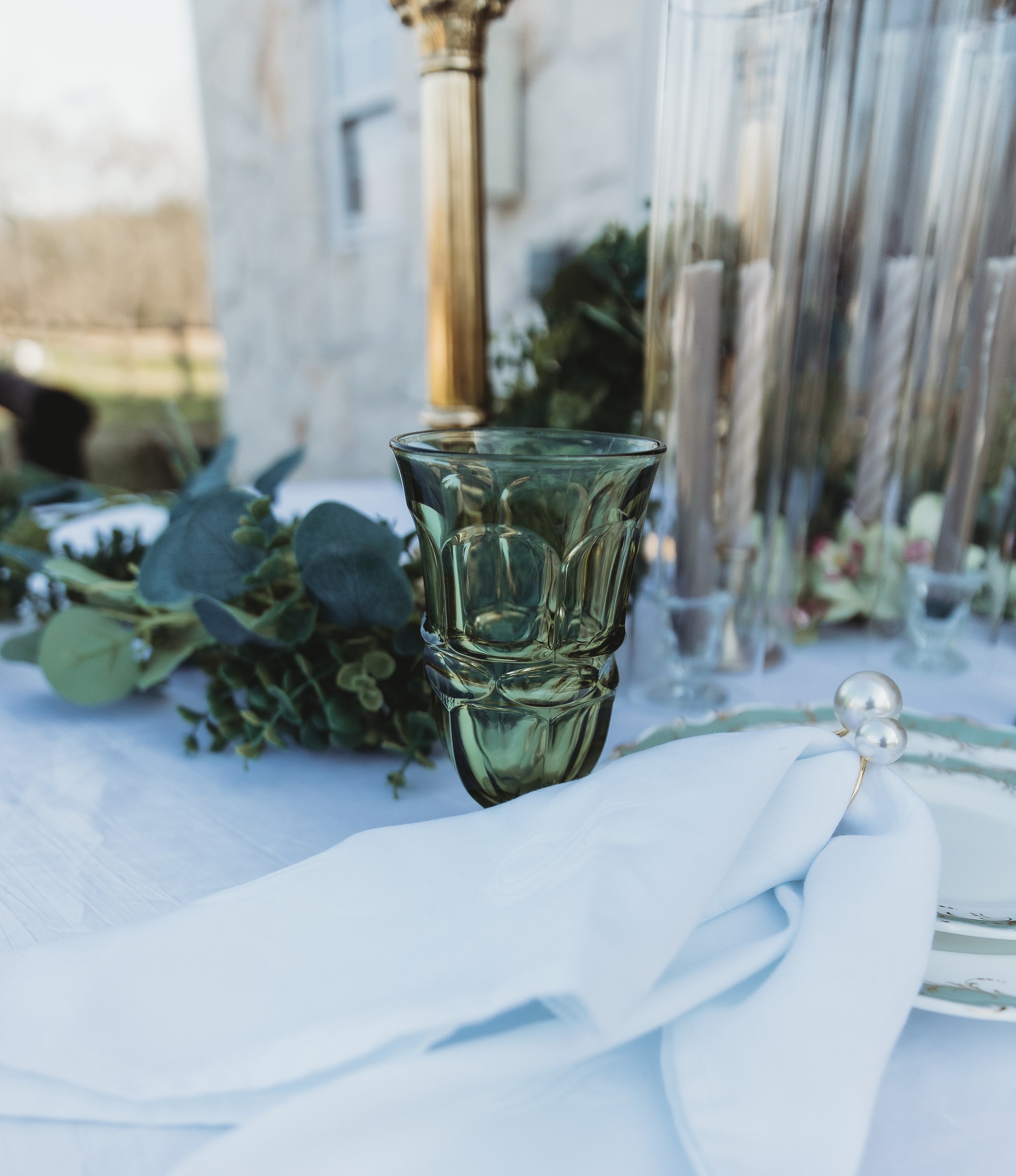 A clear green glass vase on a white tablecloth with a white napkin and pearl-topped napkin ring, greenery to the left, and tall candles in glass holders in the background.