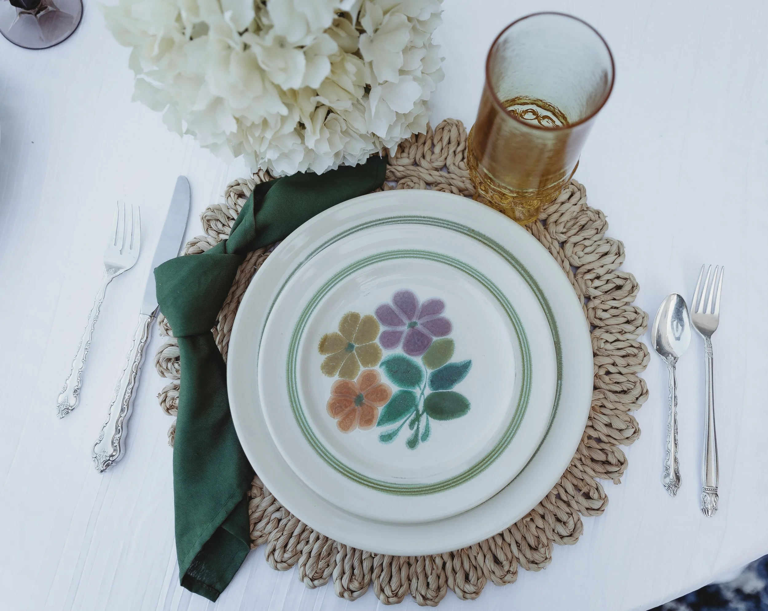 A table setting with a decorative floral plate, a green cloth napkin, a glass of beverage, a bouquet of white flowers, and silverware on a round woven placemat.