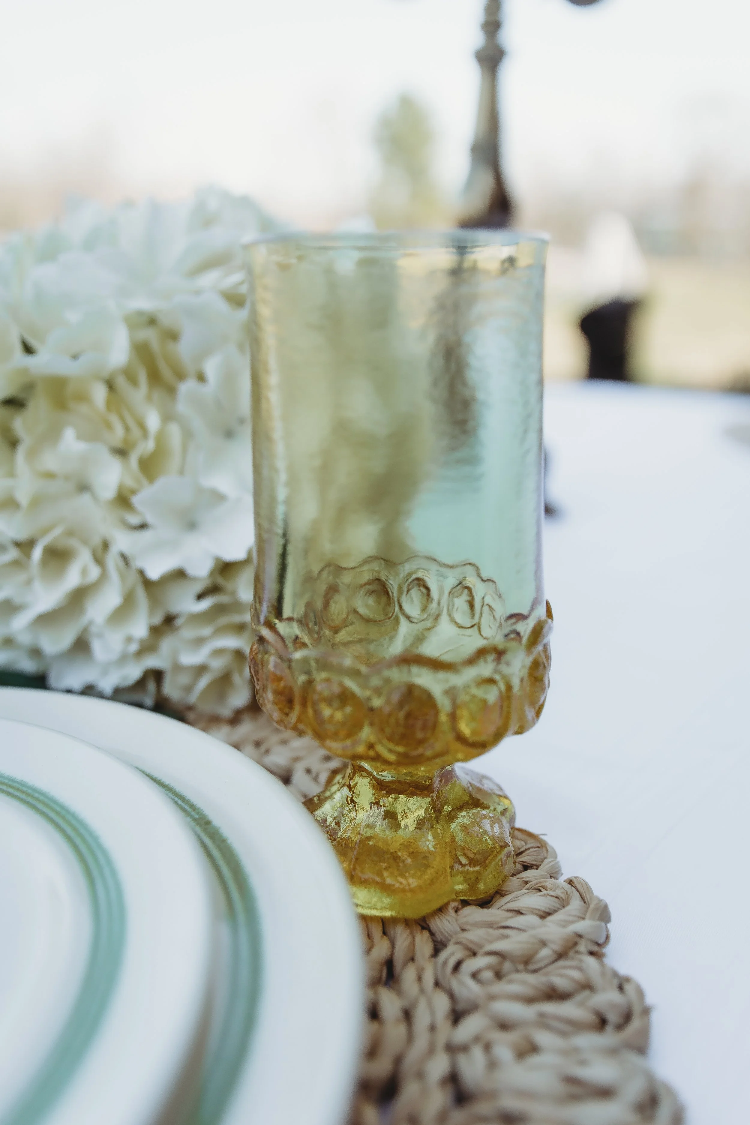 Close-up of a yellow glass goblet on a woven placemat with a white floral arrangement in the background, set on a dining table.