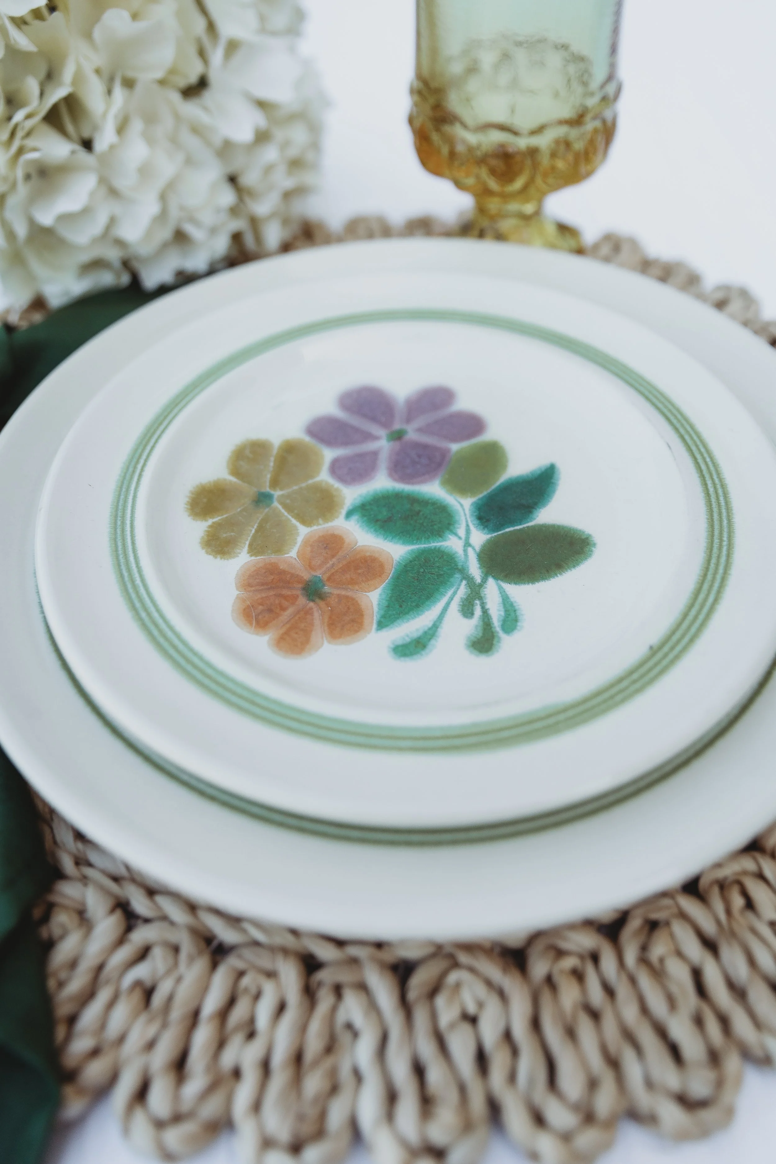 Decorative plate with colorful floral pattern, set on a woven placemat with flowers and a candle in the background.