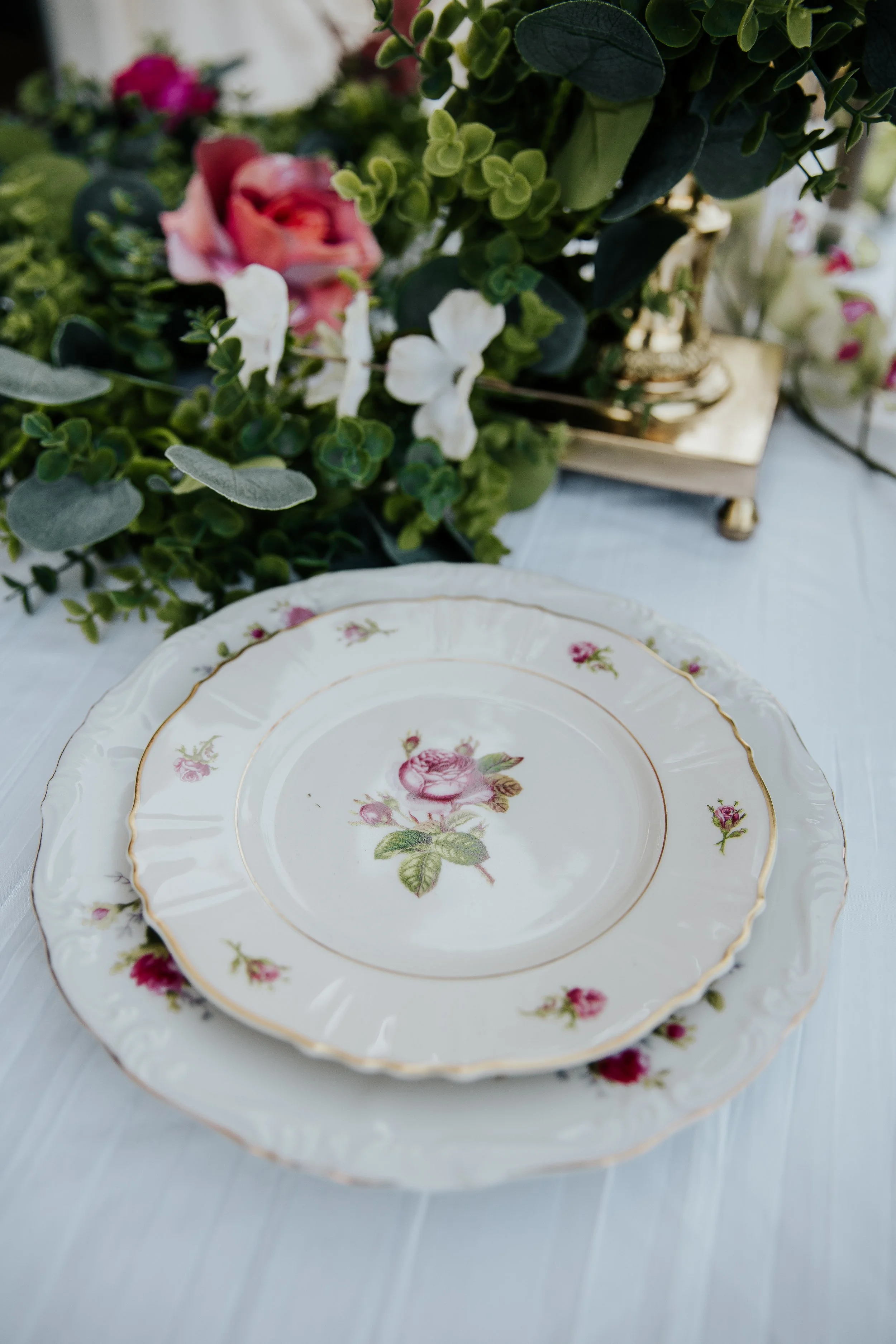 Stacked elegant floral patterned china plates with pink roses and green leaves, set on a white surface with a floral arrangement in the background.