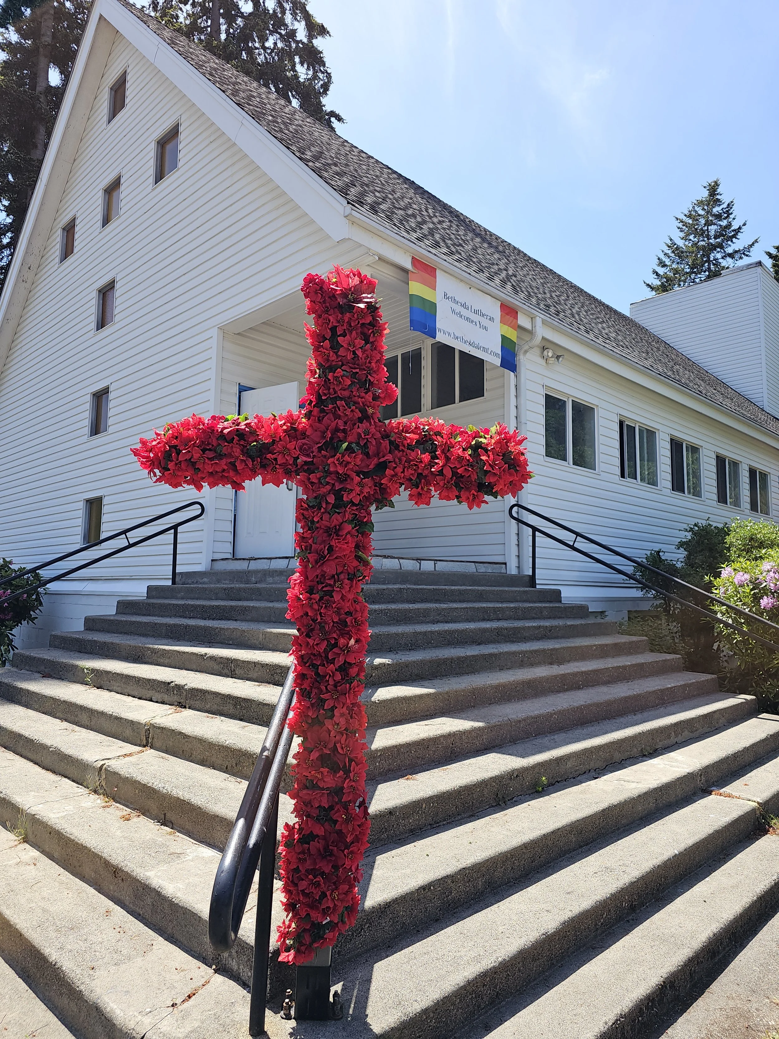 Cross wrapped with red flowers for Pentecost in front of a traditional church building with a pride flag above the entrance