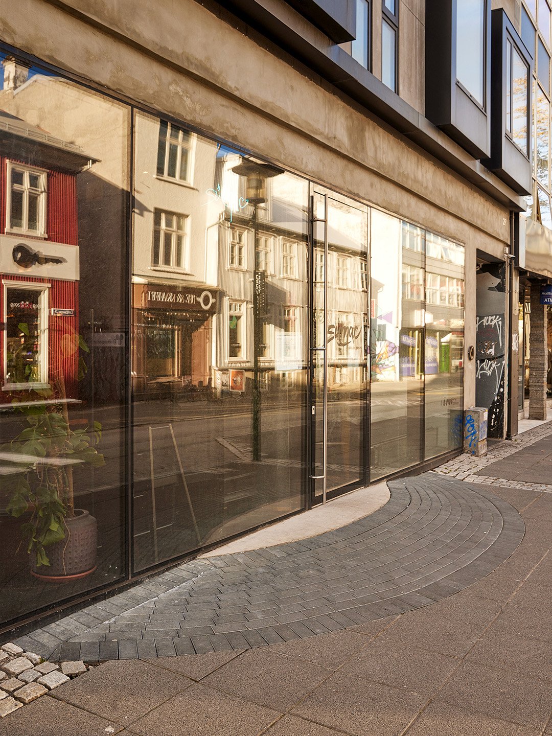 Street view with modern building featuring large glass windows reflecting older buildings, sidewalk with curved paving, and minimal graffiti on a wall.