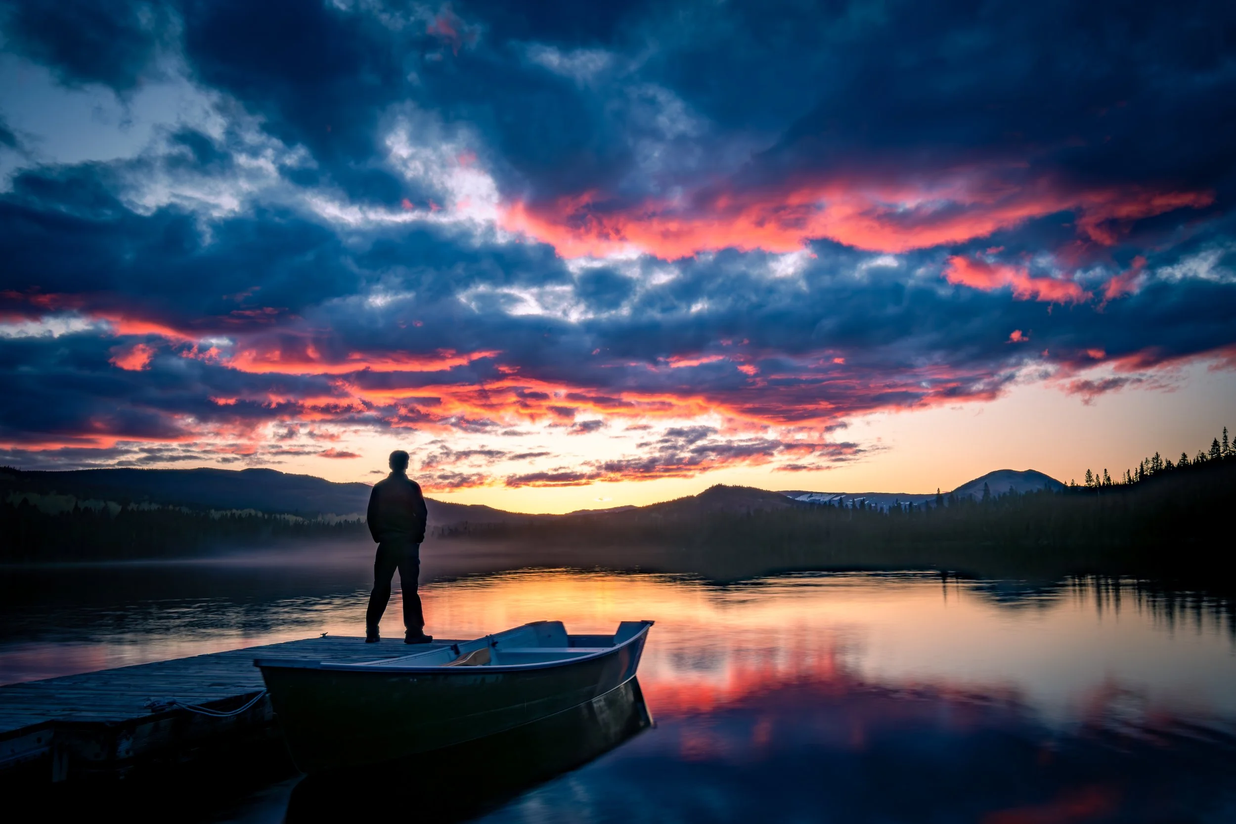 Silhouette d'une personne debout sur un quai au bord de l'eau, regardant un coucher de soleil avec des nuages colorés dans le ciel, entouré de montagnes et d'arbres.
