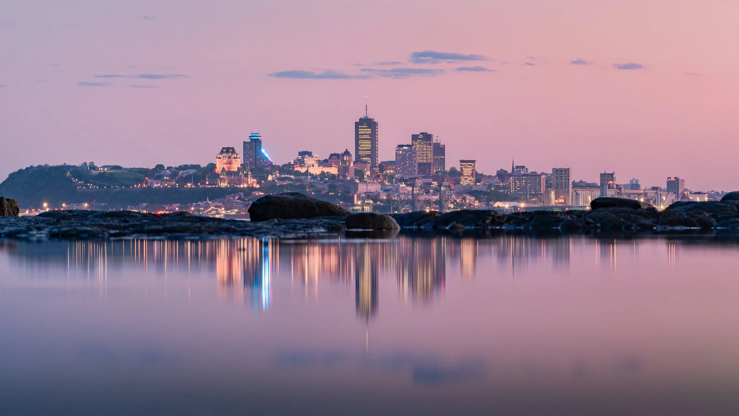 Vue de la ville de nuit avec un reflet dans l'eau calme, ciel pastel, gratte-ciel illuminés et montagnes en arrière-plan.