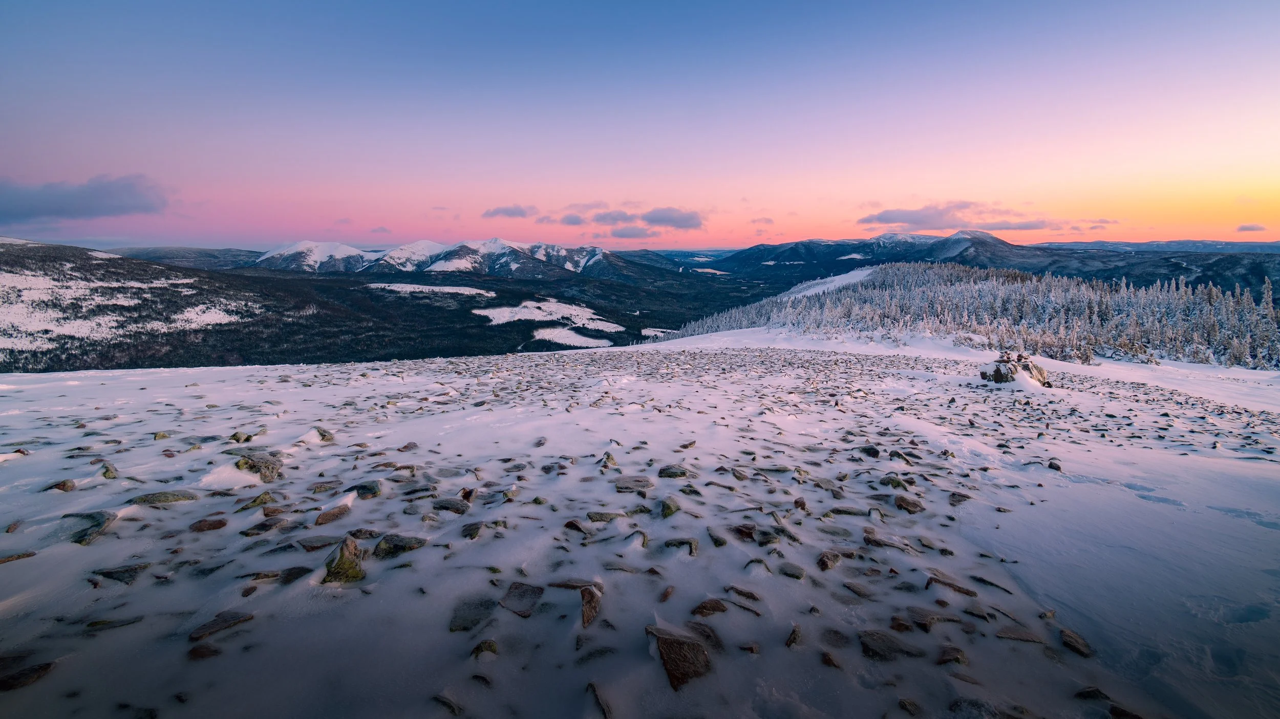 Paysage de montagne enneigée avec un ciel coloré au coucher du soleil, en avant-plan des pierres recouvertes de neige, forêts et montagnes lointaines.