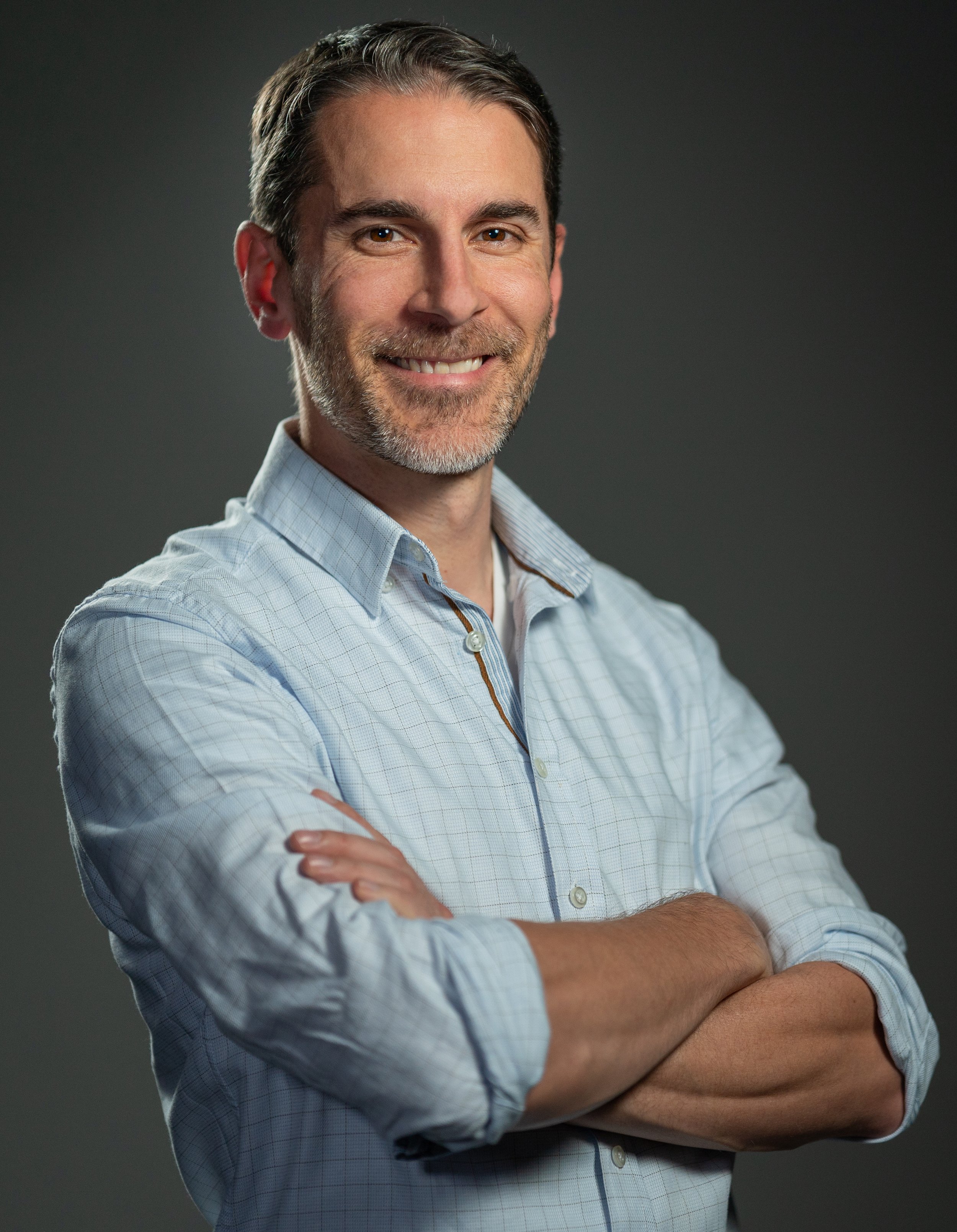 Homme souriant avec des bras croisés, portant une chemise à carreaux claire, contre un fond gris
