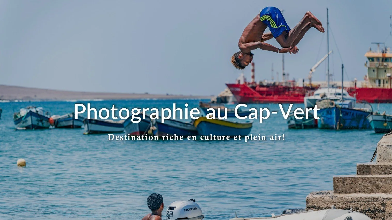 Un jeune homme en plein saut dans l'eau, avec des bateaux en arrière-plan au Cap-Vert.