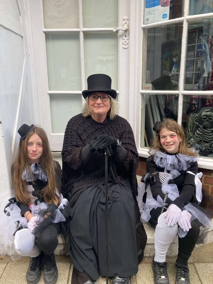 Three people dressed in Victorian costumes sitting in front of a house. The woman in the middle is wearing a black top hat, glasses, a black shawl, and holding a cane. The two girls on either side are dressed in black and white costumes with ruffled collars, gloves, and small black top hats.