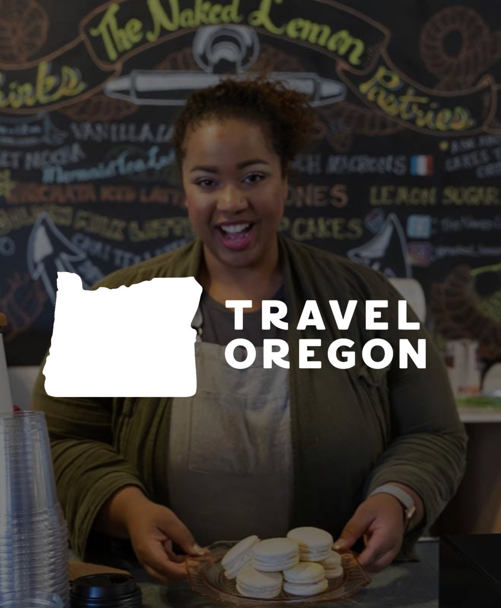 Person smiling while holding a plate of macarons in a pastry shop with "Travel Oregon" text overlay.