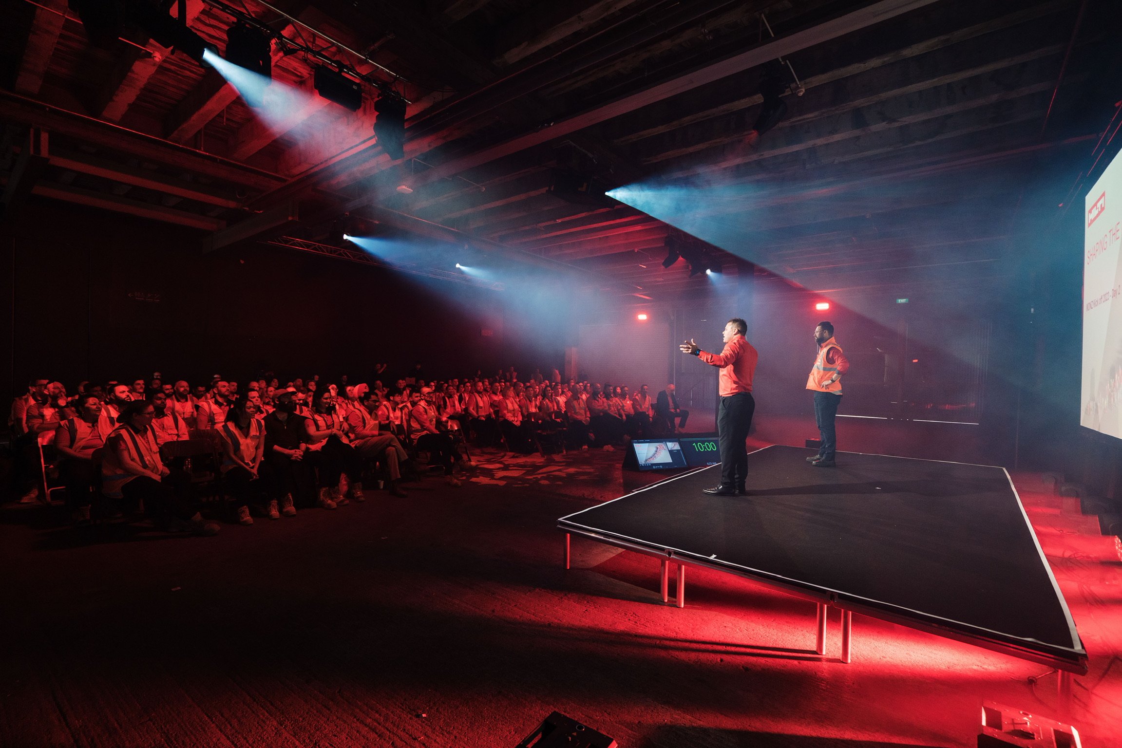 Conference speakers on stage addressing a seated audience in a dimly lit venue with red lighting and spotlights.