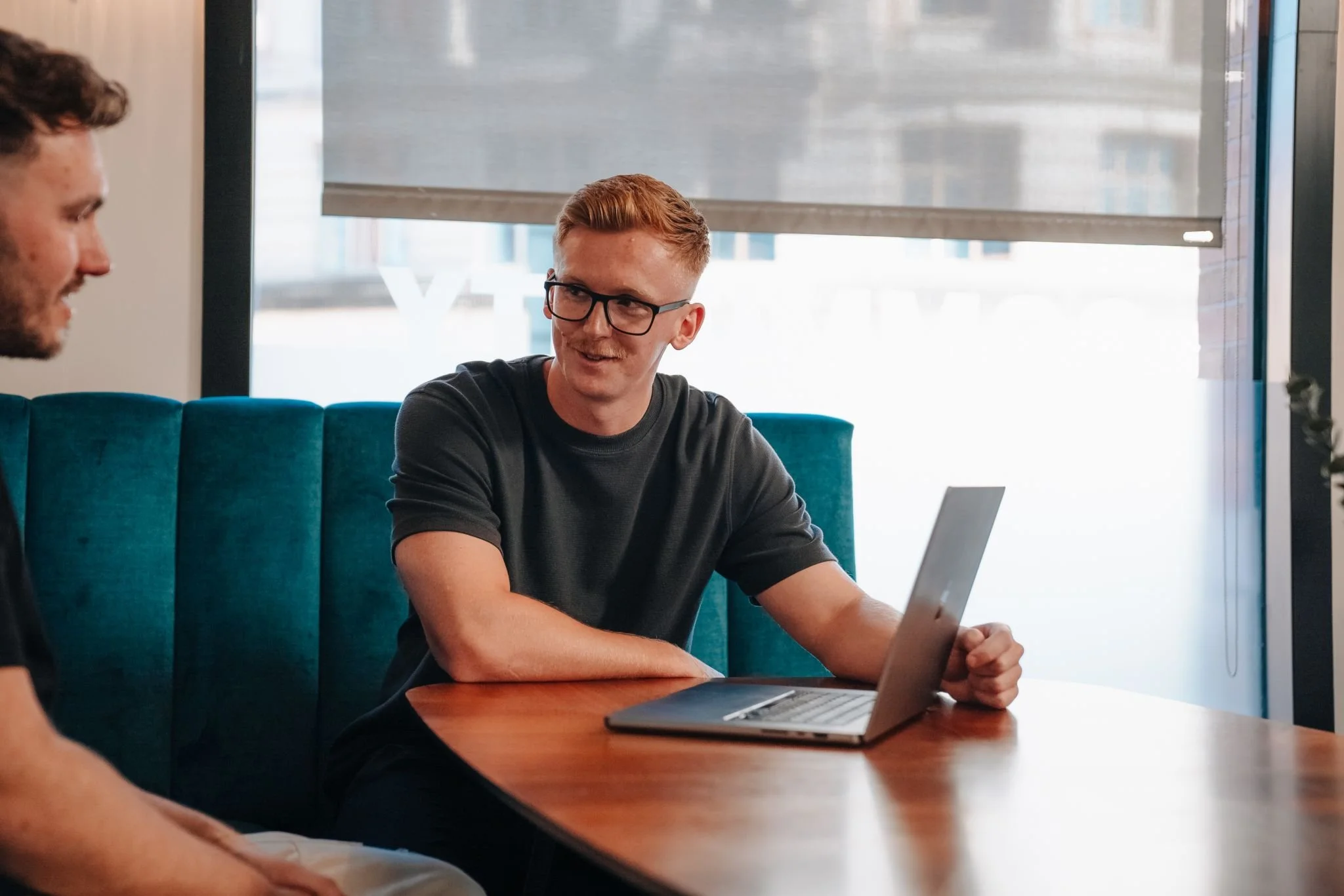 Two men sitting at a wooden table, one with glasses working on a laptop, having a conversation in a modern office with large windows and teal seating.