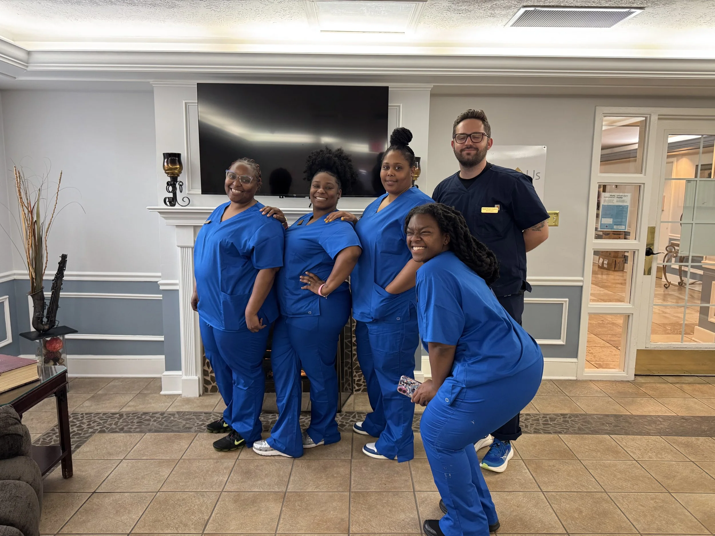 Group of five healthcare workers in blue scrubs posing indoors, with one man in black scrubs, standing in front of a flat-screen TV mounted on a white wall, in a room with decorative wall moldings and a glass double door to the right.