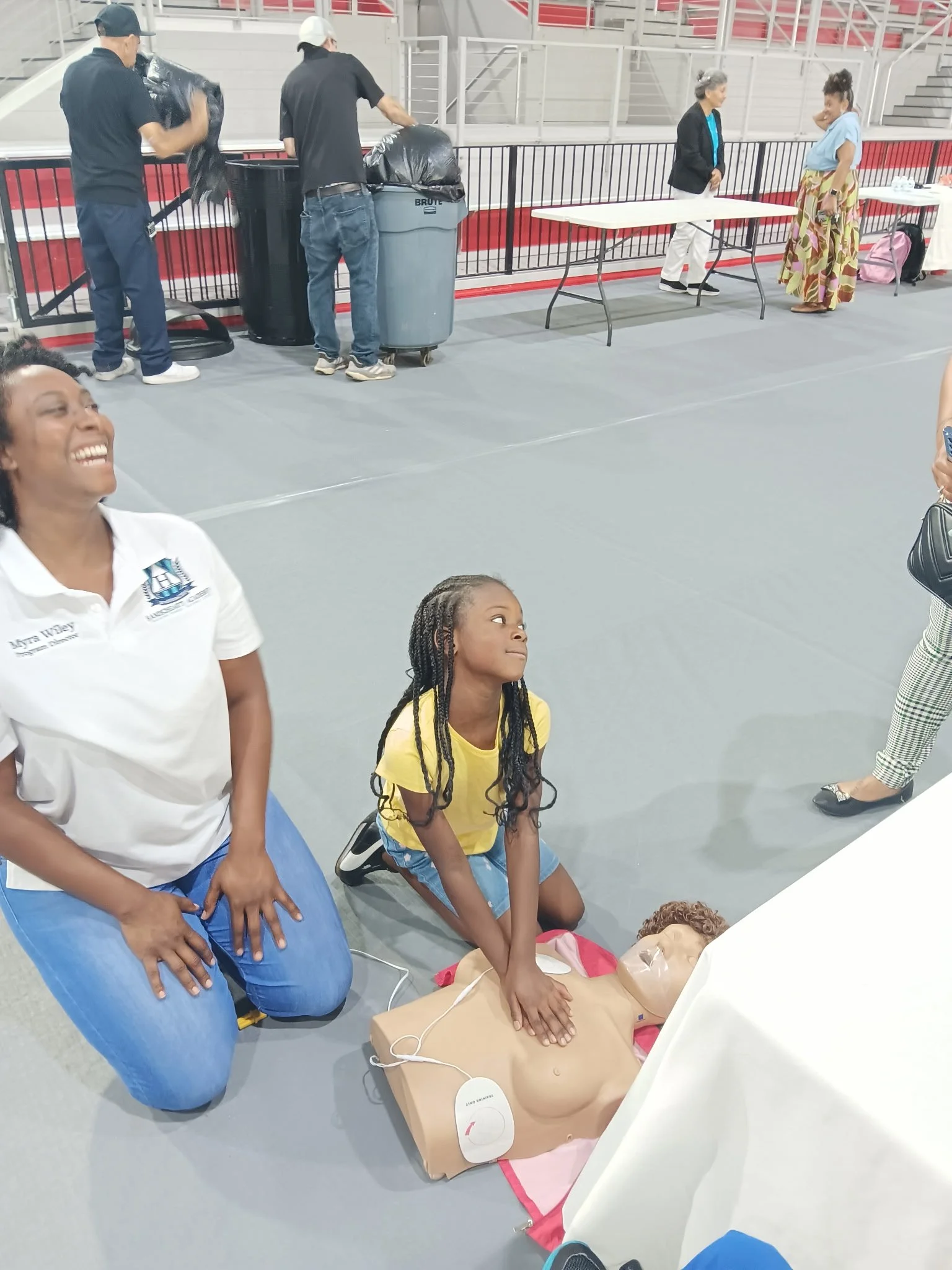 Group of people practicing CPR on a mannequin on a table, with others watching in a gymnasium or event space.