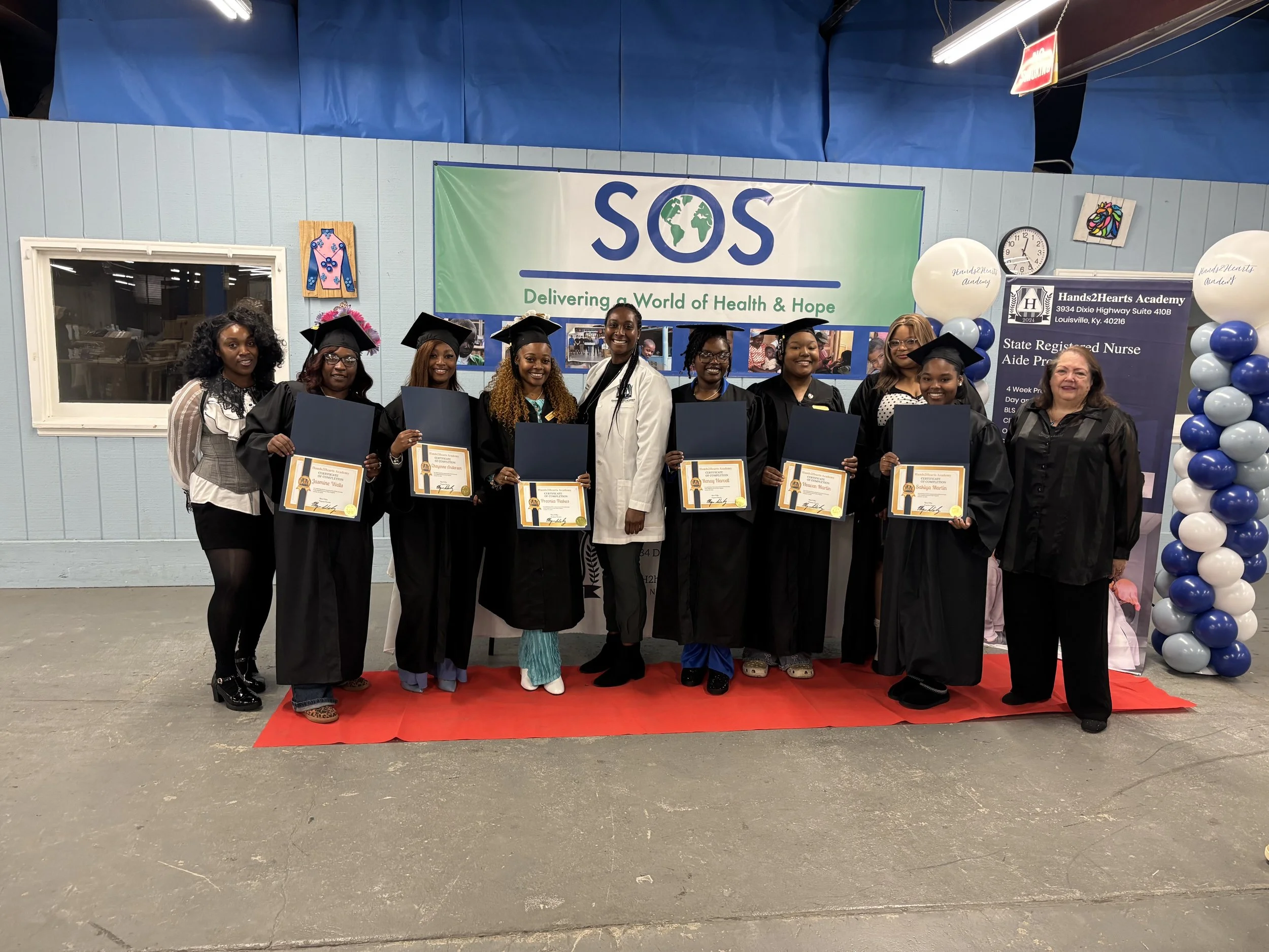 Group of women in graduation regalia holding diplomas, standing on a red carpet in front of a backdrop with SOS and globe logo, balloons, and event information at Hands2Hearts Academy.