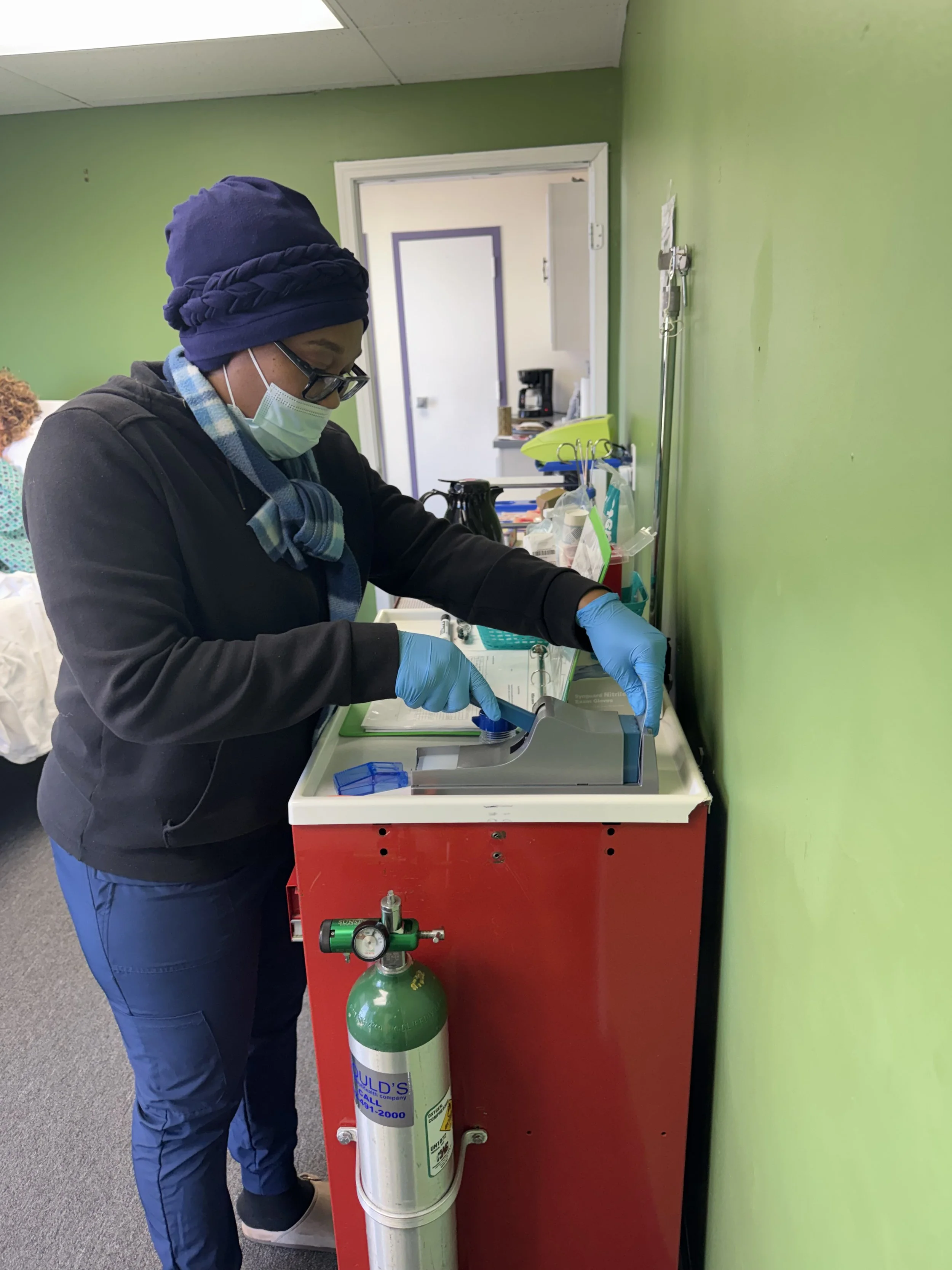 A person wearing a navy blue head covering, glasses, a face mask, and blue gloves working at a medical station with supplies, a green oxygen tank, and a red cart against a green wall in a healthcare setting.