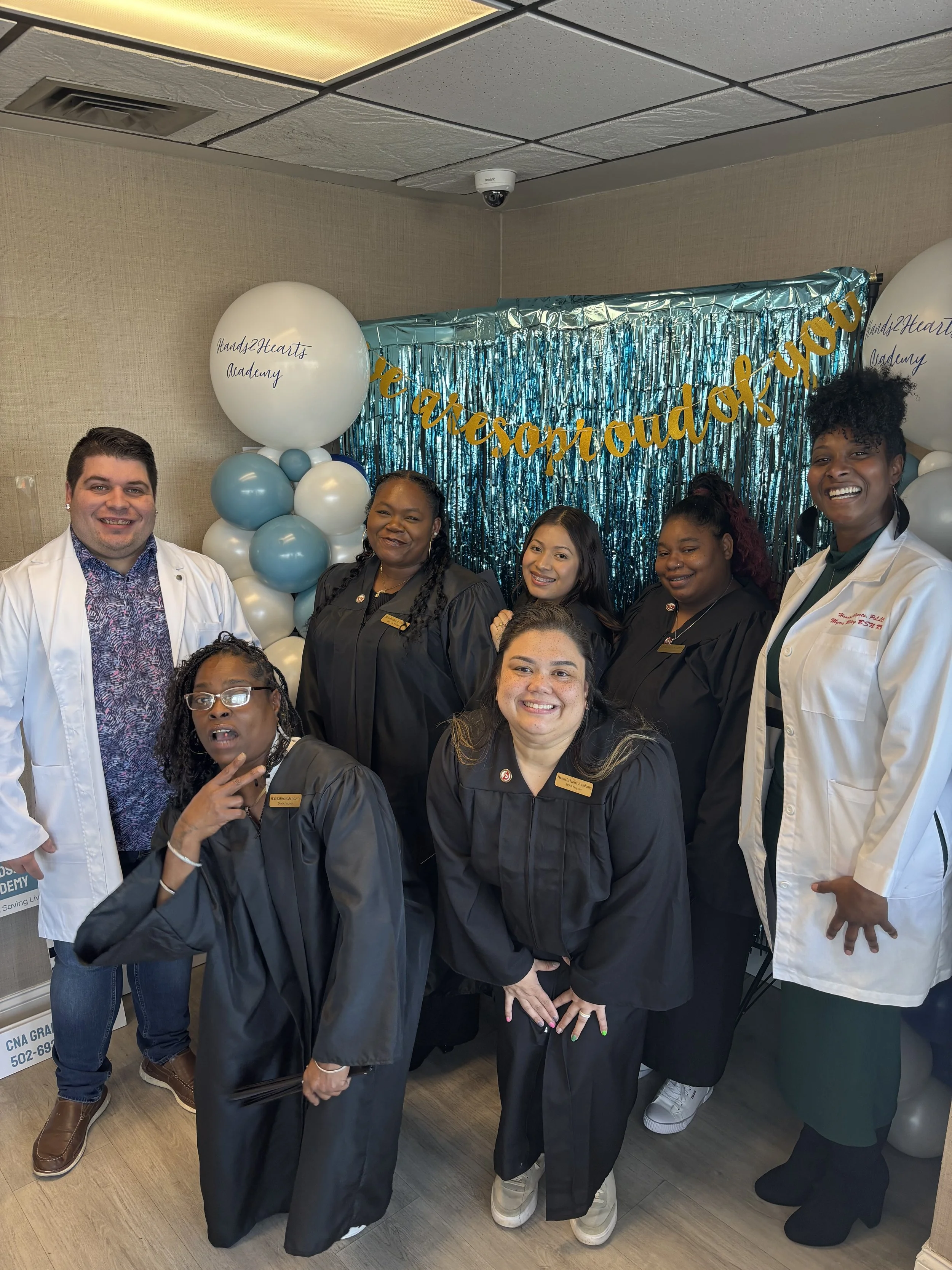 A group of diverse smiling healthcare professionals, including doctors and nurses, in front of a blue foil birthday backdrop with balloons that say 'Hands2Hearts Academy' and a banner that says 'You are outstanding'.